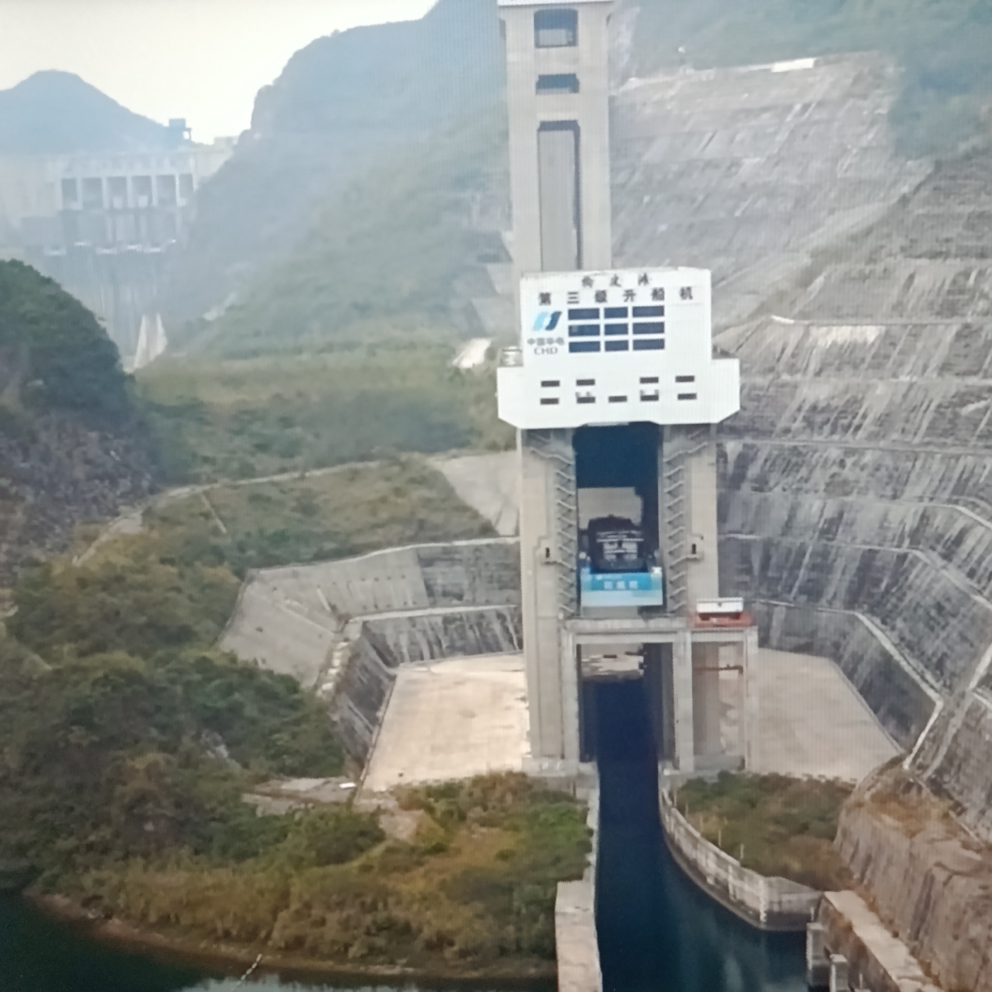 The image shows a large concrete dam and a vertical ship lift. The dam is situated in a hilly, green landscape. The ship lift is a prominent structure with a tall tower and a large elevator car. The elevator car is visible within the shaft of the lift, appearing to be a substantial piece of machinery. The overall scene suggests a large-scale civil engineering project, likely for hydroelectric power generation or flood control. There are no people visible in the image. The location is identified as the city of Dubăsari town council, Moldova.
