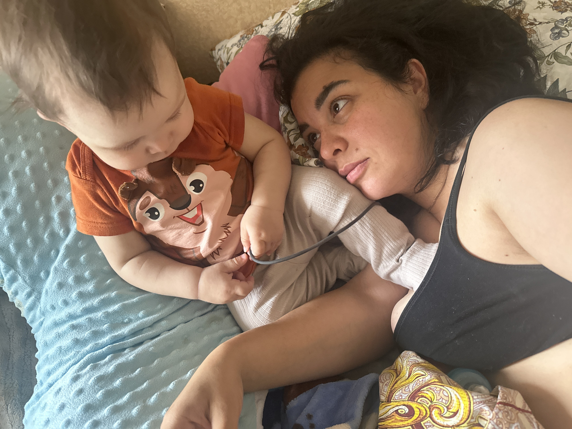 This media file shows a close-up, high-angle shot of a woman and a baby lying down together. The woman has dark, curly hair and is wearing a black tank top. She is looking towards the baby, with a pensive expression on her face. The baby, facing away from the camera and towards the woman, has brown hair and is wearing an orange t-shirt with a cartoon character on it. The baby's hands are visible, and they appear to be holding onto something thin and grey, possibly a cord. They are lying on a blue, textured blanket. In the background, there are pillows, one of which is pink, and another with a floral pattern. The overall scene suggests a moment of quiet intimacy between the mother and child. The lighting suggests it might be daytime, but there are no strong indicators of the specific time or weather. The setting appears to be indoors, possibly a bedroom or living room.