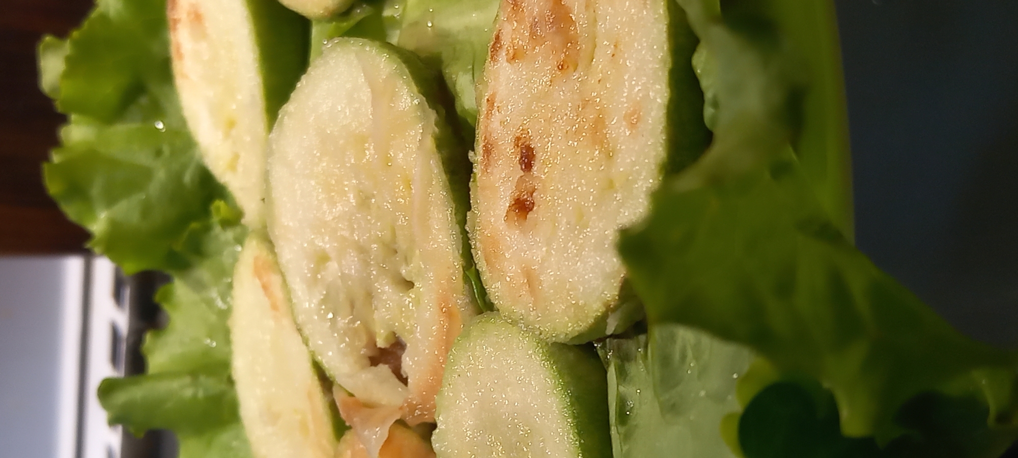The image displays several oval slices of green-skinned fruit, consistent with guava, arranged on a bed of vibrant green lettuce leaves. The fruit's interior is pale yellow to off-white, with some slices exhibiting localized brown spots and a surface covered in small, glittering particles, indicative of sugar or moisture. The lettuce leaves beneath appear wet. The blurred background reveals horizontal white lines in the upper left, an indistinct dark brown object in the upper right, and a dark, undifferentiated surface in the lower right, suggesting an indoor setting. This scene is situated in Kryvyi Rih, Ukraine.