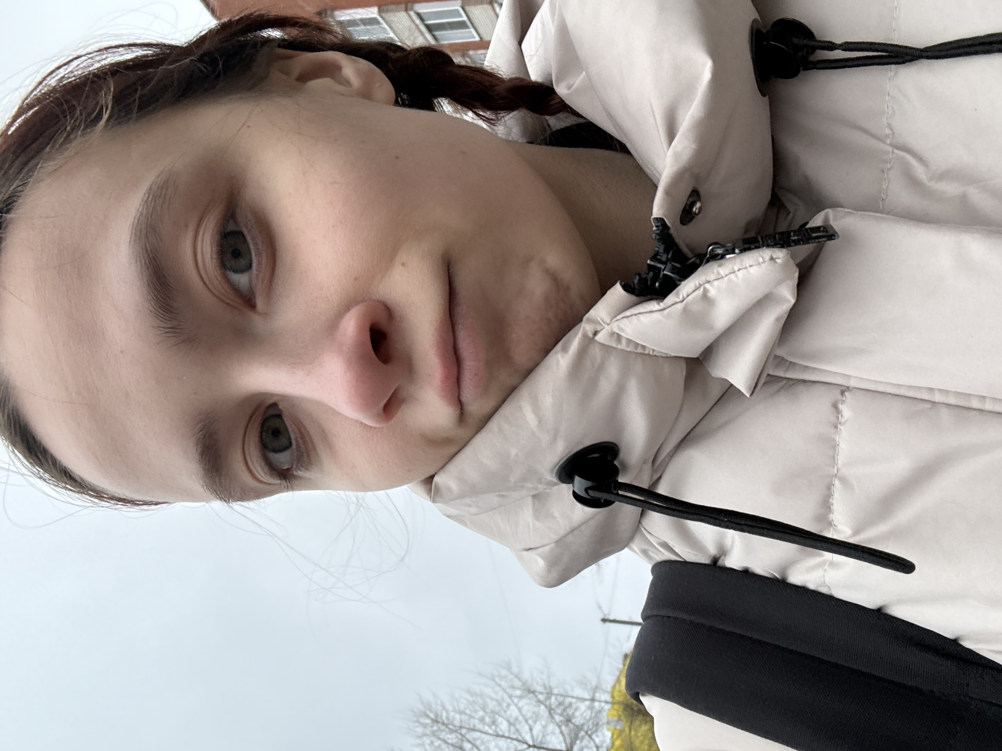 A selfie portrait of a light-skinned individual with braided brown hair, fair complexion, and visible facial features, including light eyes, nose, and lips. The person is wearing a light-colored, possibly beige, puffer jacket with black drawstrings and a zipper, which is partially visible. A black backpack strap is seen on the right side of the image.
The background shows a cloudy sky and blurred elements of a city environment, including the top of a building with windows and bare tree branches. The location is identified as the city of Chaikovsky, Russia, based on the request. No specific actions or interactions are observed, as the subject is posing for the photograph.