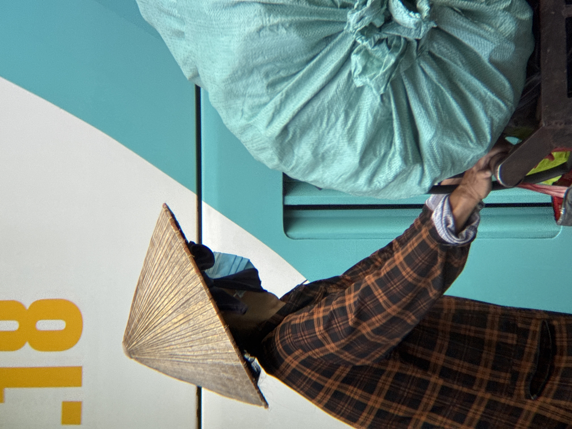The image captures a close-up view of a person wearing a conical hat and a plaid shirt, pushing a cart laden with a large, turquoise-colored bag. The person is also wearing a blue face mask. The cart appears to be in motion, as suggested by the blurred background and the dynamic pose of the person.
The background is divided by a diagonal line, with the upper left section featuring a white surface with large yellow numbers "18" and a dot. The lower right section is a bright turquoise color, possibly indicating the side of a vehicle or building. The overall scene suggests an urban environment, likely in Nha Trang, Vietnam, given the presence of the conical hat, a common headwear in Southeast Asia, and the style of the cart.
The focus is on the person and the cargo, with the background elements providing context. The lighting suggests it is daytime. The composition is tight, emphasizing the texture of the hat, shirt, and the crinkled surface of the bag. The person's hand is visible on the handle of the cart, indicating movement and action.