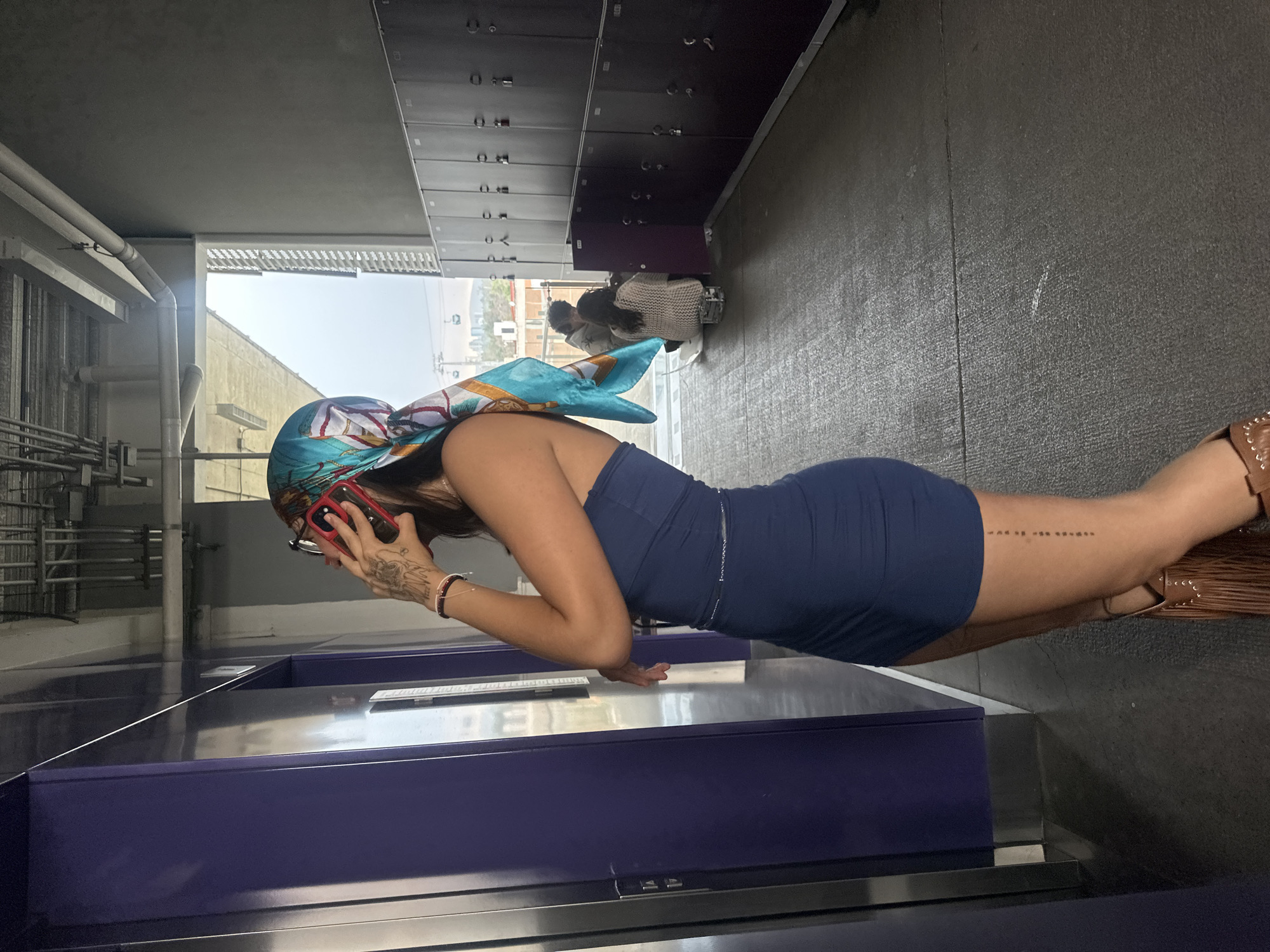 A woman wearing a blue dress and a patterned headscarf is standing next to a table in what appears to be a public space, possibly a bus or train station in Mexico City. She is holding a red smartphone to her ear with her right hand, and her left hand is resting on the edge of the table. The table is purple and has a stainless-steel trim. Behind her are rows of metal lockers. In the background, through a window, there is a view of buildings and a blue sky, suggesting an urban environment. Two other people stand on a walkway in the background. The floor is a dark grey or black.