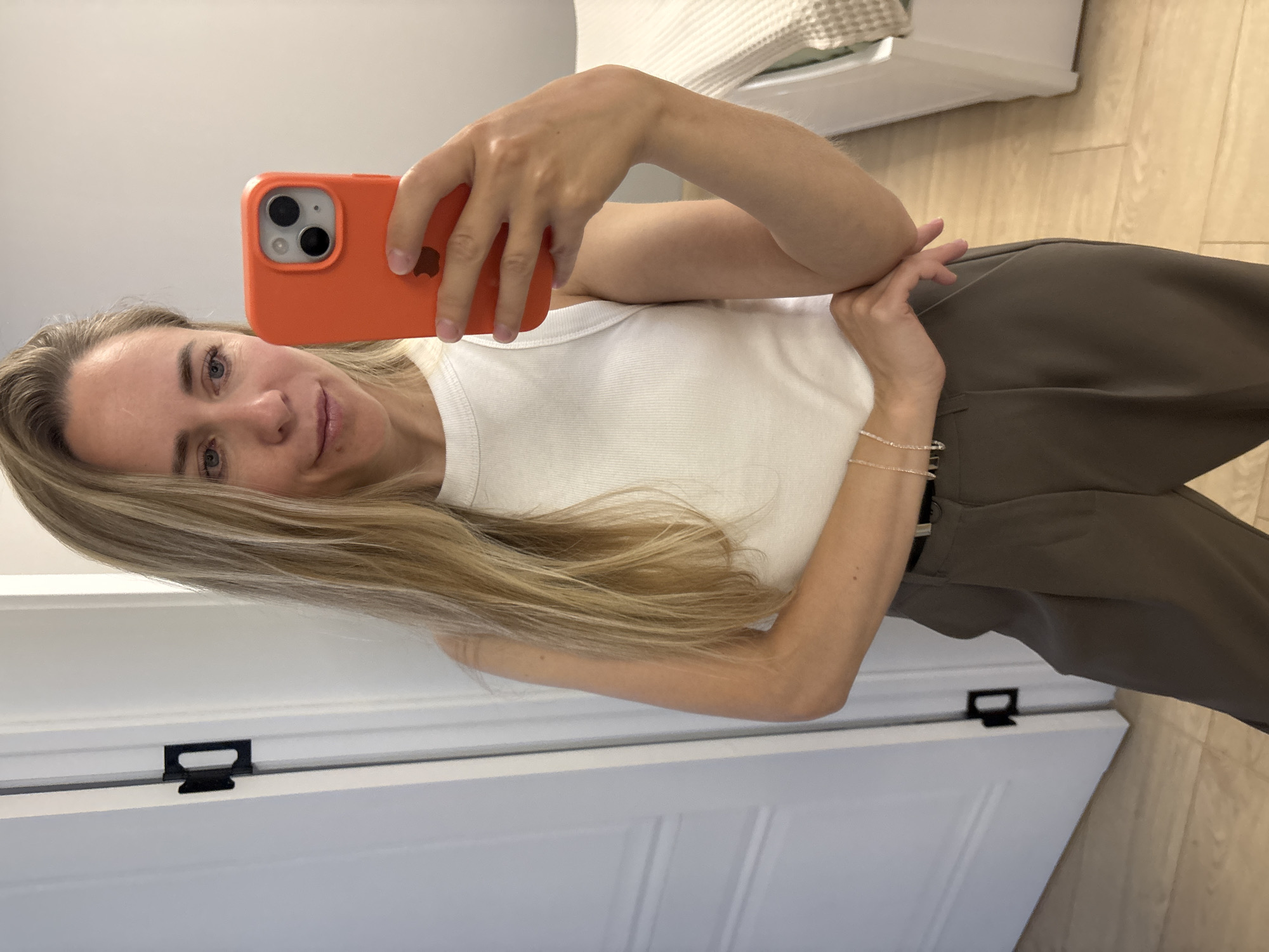 A blonde woman with long hair is taking a selfie in a room. She is wearing a white ribbed tank top and dark brown pleated pants with a black belt. She is holding an orange iPhone in her right hand. Her left arm is bent, with her hand resting on her hip. The background shows a light-colored wall and a door with black hardware. Part of a bed with a textured blanket is visible on the right side of the image. The floor is made of light-colored wood planks. The image appears to be taken indoors, possibly in a home. There is no discernible information in the image that can pinpoint the location to Chaikovsky, Russia.