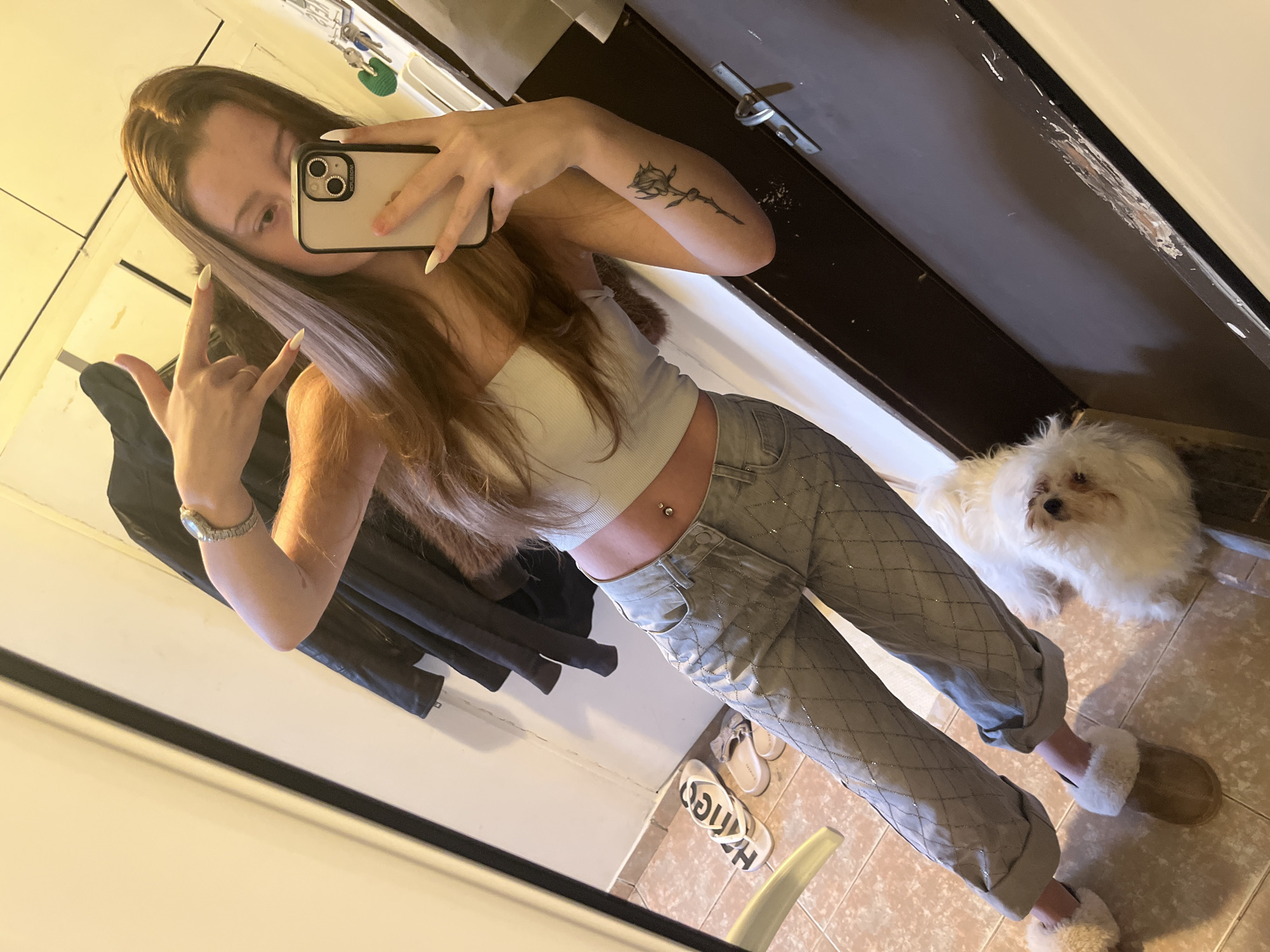 A young woman with long brown hair and a light-colored top is taking a selfie in a room. She is wearing light grey pants with a quilted pattern and slippers. She is making a hand gesture with one hand and holding a phone in the other. A small white dog is sitting on the floor near her feet. There is a dark-colored door in the background, along with a coat rack and other items. The room appears to be a hallway or entryway with tiled flooring. There is no evidence to support the scene being located in Varna, Bulgaria.