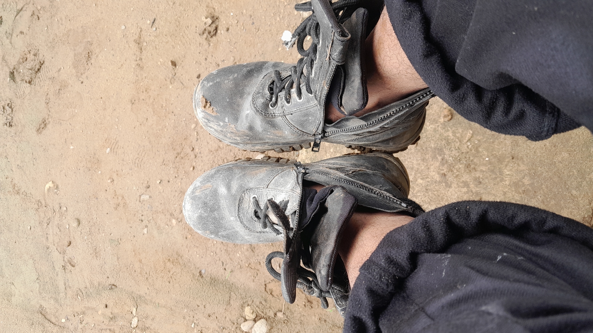 A top-down view shows a person wearing black, weathered boots with zippers partially open and laced. The boots are standing on a sandy surface. The person is wearing black pants, partially obscuring the view of the boots and ground. The location is Lhokjok, Indonesia.