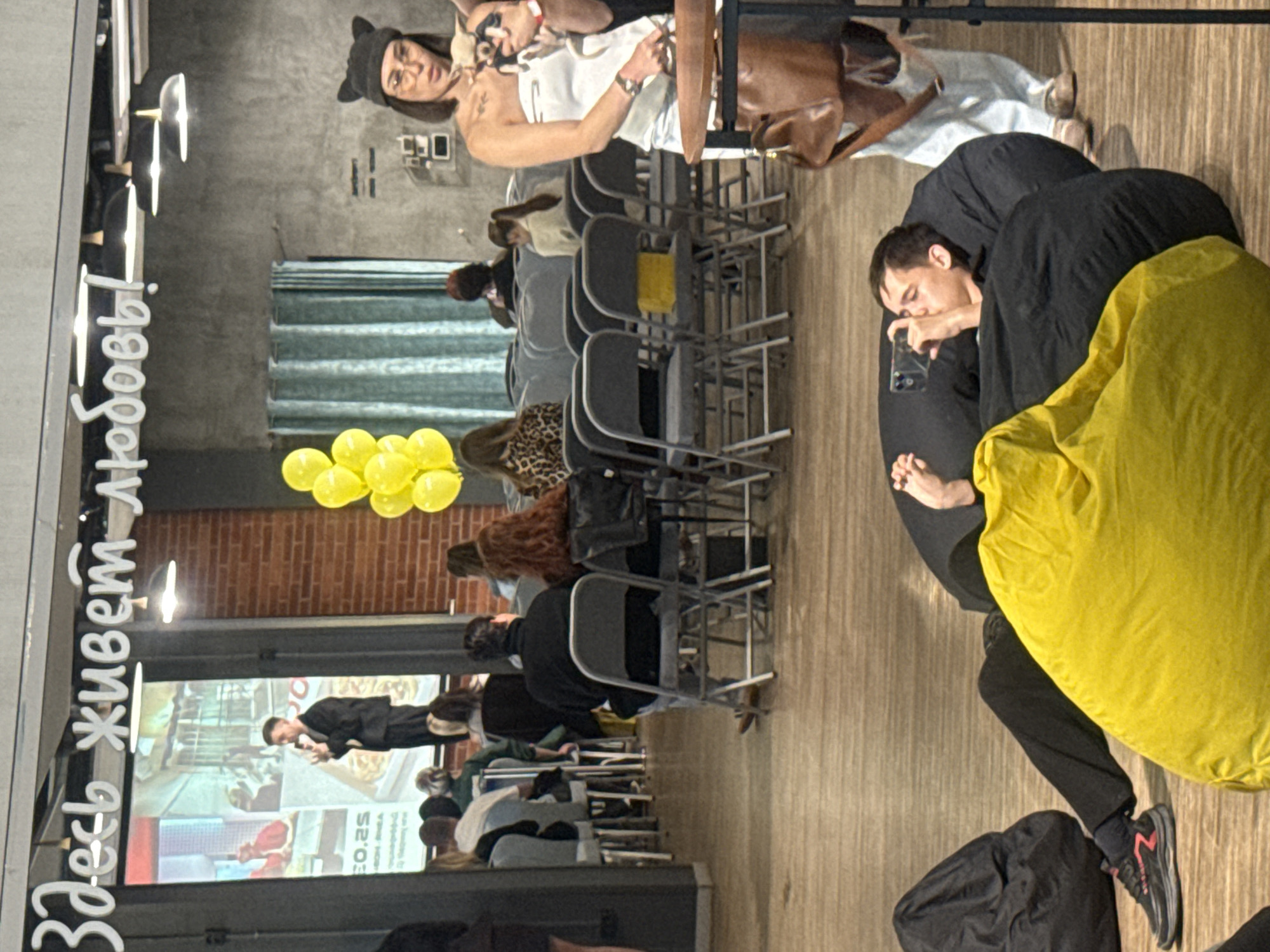 This indoor image captures an event in Vladivostok, Russia, identified by the overhead Cyrillic text "Здесь живет любовь!" which translates to "Love lives here!" The scene features a speaker on a stage at the left, addressing an audience. The speaker, a man dressed in a dark suit, holds a microphone and stands before a large projector screen displaying images and some Cyrillic text, including "EO.2S" and partial words.
The audience is seated on rows of dark-framed chairs with light grey upholstery, facing the stage. Many individuals are visible from behind, observing the speaker. In the foreground, on the wooden floor, two beanbag chairs are present: one yellow and one black. A young man is reclining on the black beanbag chair, holding a black smartphone to his eye, appearing to record or photograph the event. He wears dark pants and black and red sneakers.
To the right, a woman stands with a small, light-colored dog in her arms, which she holds near a small wooden table. She wears a strapless white top, light blue jeans, eyeglasses, and a black beanie with cat ears. A brown leather shoulder bag rests on the table next to her. Yellow balloons are clustered on the floor to the right of the stage.
The environment includes concrete walls, one section featuring exposed red brick, and another section with a light blue striped curtain. Black pendant lights hang from the ceiling. The overall impression is a casual, contemporary event space.