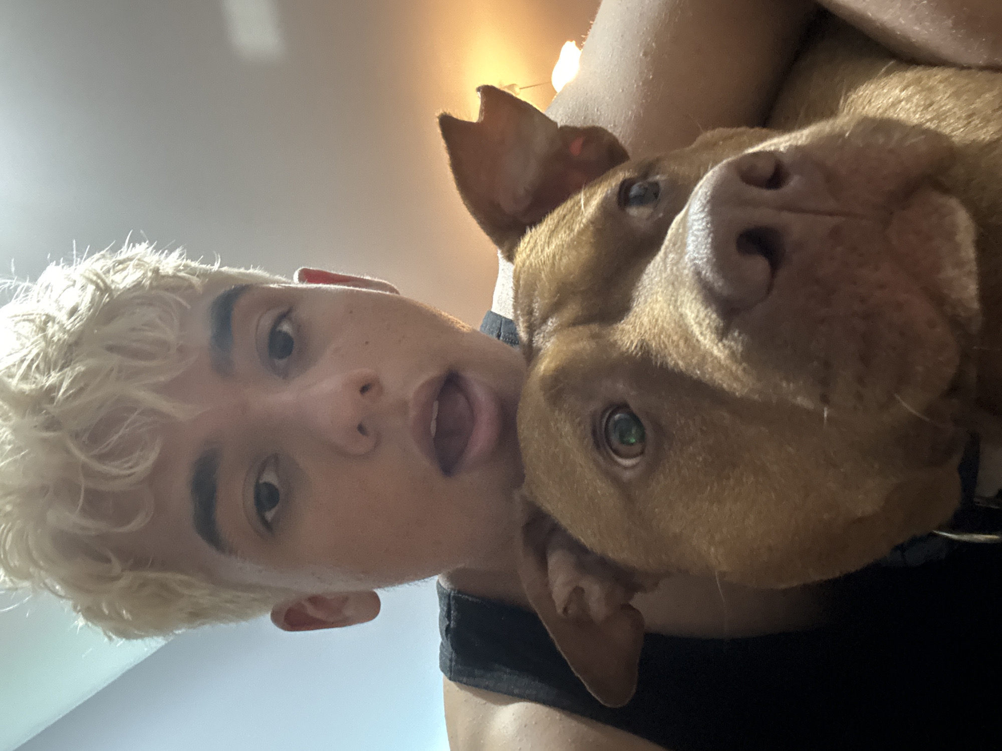 The image is a close-up selfie featuring a young person with bleached blonde, curly hair and a pit bull dog. The person is wearing a black tank top and is looking directly at the camera with their mouth slightly open. The dog, which is a reddish-brown color, is positioned in front of the person, with its face filling a significant portion of the frame. The dog's head is tilted, and its eyes are looking to the side. The background is out of focus, showing a gradient of light blue and grey, suggesting an indoor environment with artificial lighting. There are no visible landmarks that can be used to determine the location in the city of Sofia, Bulgaria.