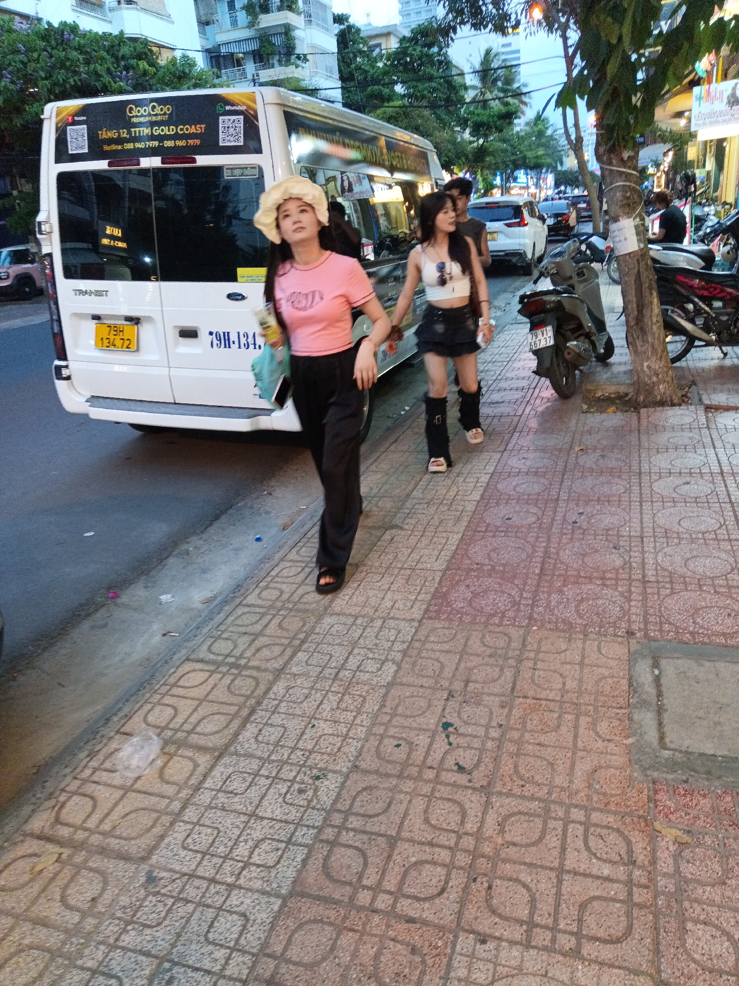 This image was taken on the street in Nha Trang, Vietnam. In the foreground, two young women are walking along a tiled sidewalk. The woman on the left wears a pink t-shirt, black pants, and a wide-brimmed straw hat. She holds a drink in her right hand and a turquoise bag in her left. The woman on the right wears a white crop top, a black skirt, and black leg warmers. She is also wearing sunglasses on her head. Behind them, a white bus is parked on the street, with Vietnamese license plates visible. Several motorcycles are parked on the sidewalk and street. Buildings and trees line the street. The lighting suggests it is late afternoon or early evening.