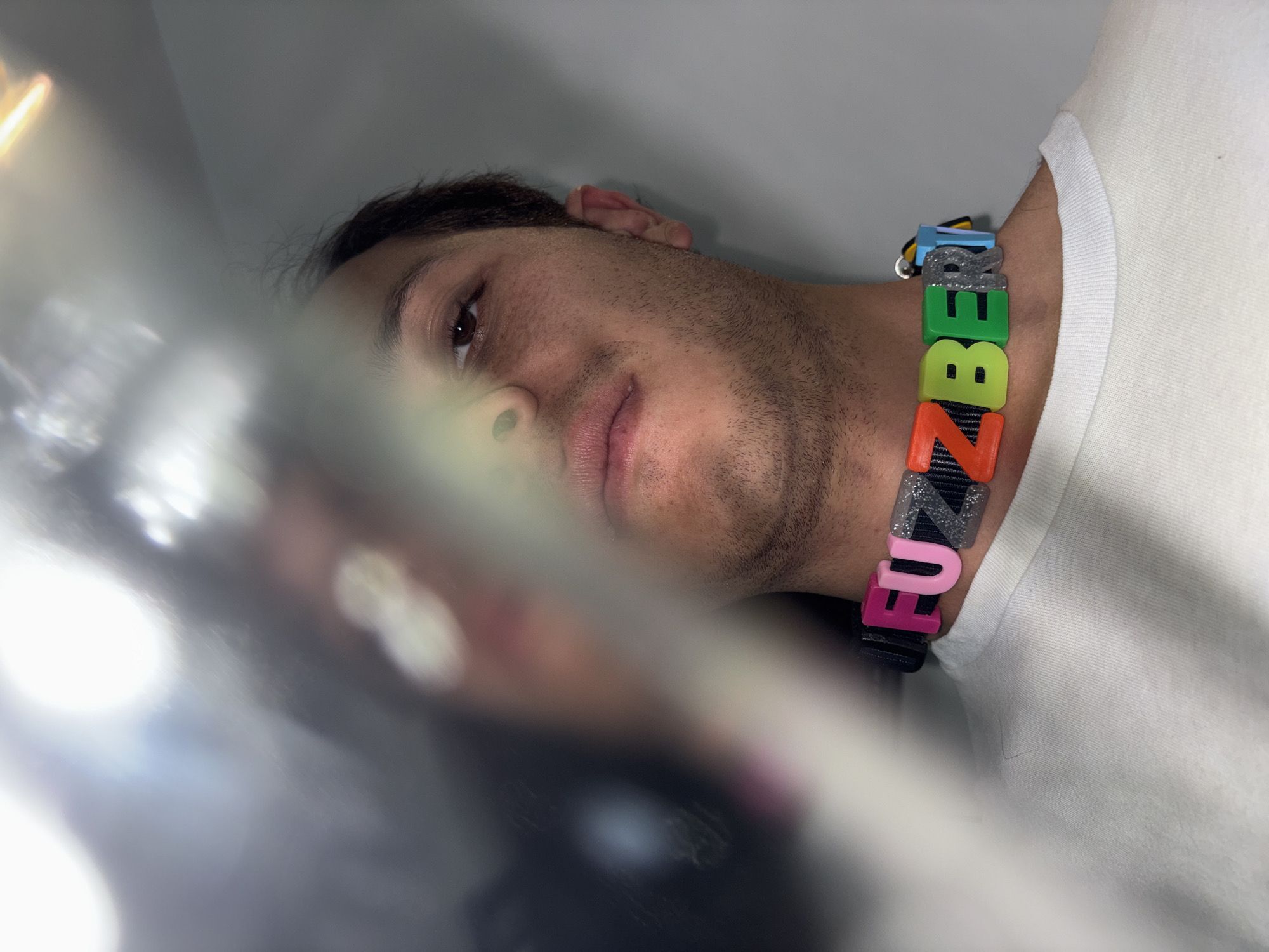A medium shot of a man with brown hair and eyes, wearing a white t-shirt and a colorful necklace or bracelet that spells out "FUNZBE," is partially obscured by a blurry, light-filled foreground element. The man's face is angled, showing his nose, lips, and right eye, with light reflecting in the eye. The bracelet is made up of individual letter blocks in blue, silver, green, yellow, orange, and pink. The location is unknown and cannot be determined from the image. The city of San Bernardino Tlaxcalancingo, Mexico is unrelated to the image.