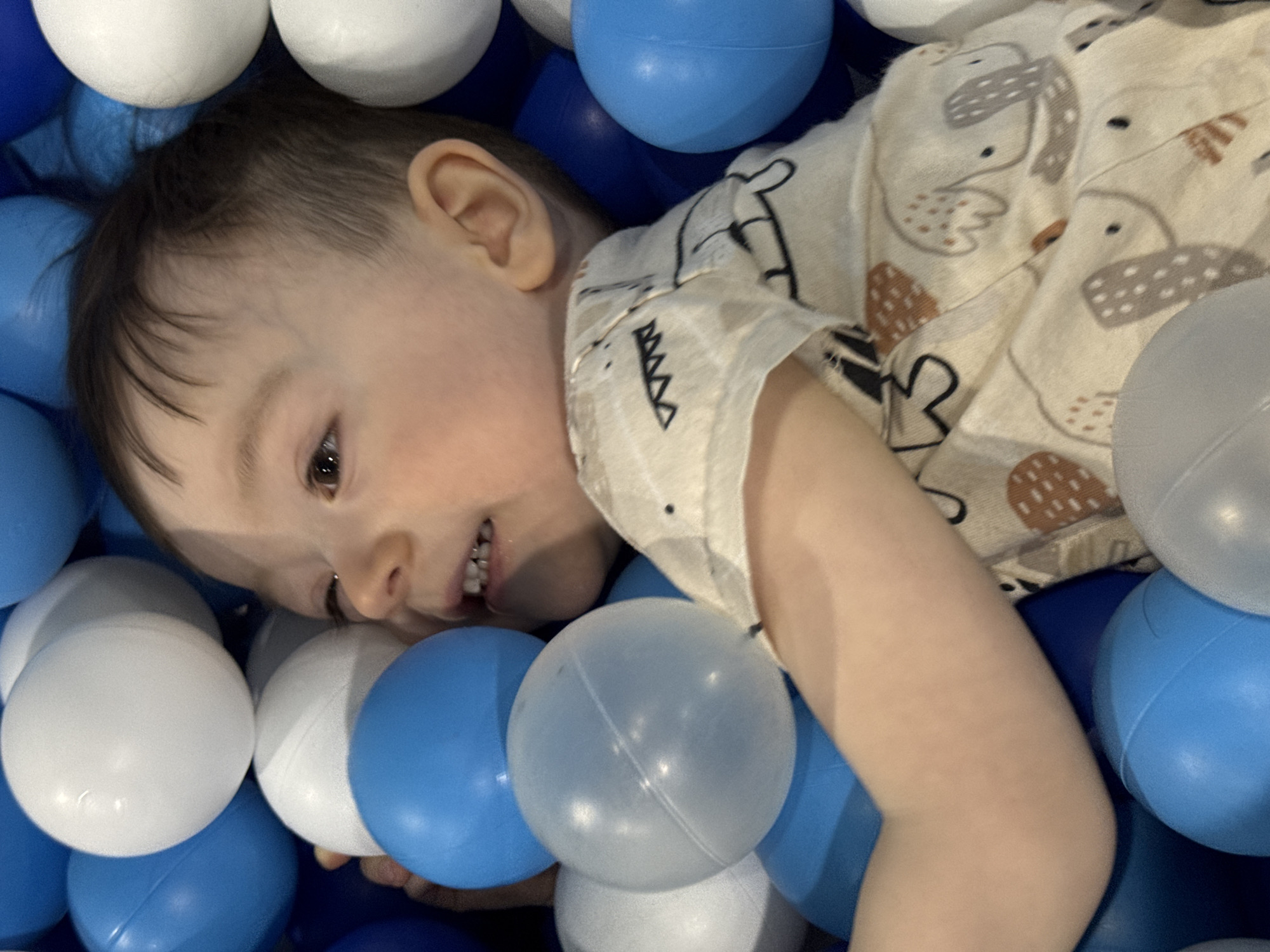 A child lies amongst blue and white plastic balls, likely in a ball pit. The child wears a patterned shirt with animal designs. The scene does not give any evidence of location in Izhevsk, Russia.