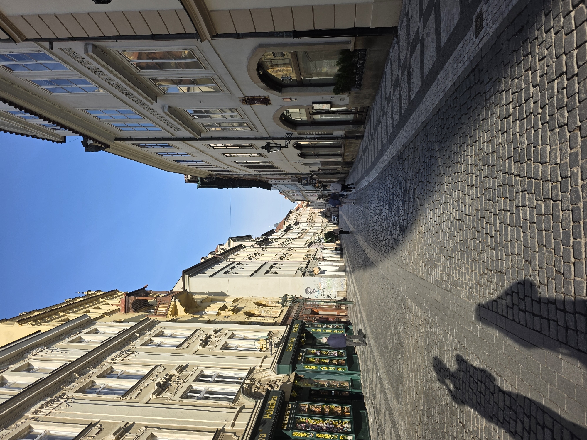 The image captures a sunlit, narrow street lined with ornate, multi-story historical buildings under a clear blue sky. The street is paved with cobblestones, and the sidewalk features a distinct checkered pattern. Long, sharp shadows stretch across the ground, indicating either morning or late afternoon sunshine.
The buildings on both sides of the street display classical architectural details, including decorative window frames, cornices, and various facade embellishments. Many windows reflect the bright sky and the opposite structures. Several shopfronts are visible, adorned with green awnings and displays.
A few pedestrians are seen walking in the distance, while one person stands closer to a shop on the right side of the street. In the foreground, two distinct shadows of people, likely the photographer and a companion, are cast prominently on the cobblestones.
Visible text on a prominent green shop awning reads "Pohádka" (Czech for "Fairy Tale"), with "PRALINKÁRNA" on a smaller sign below it. Another green awning further down shows "Czech toys" and also "Pohádka". An arched sign on a building reads "DIVADLO V CELETNÉ". Some graffiti, too indistinct to fully decipher, is visible on a building wall. The scene evokes a peaceful, everyday atmosphere in a charming, historic urban environment.