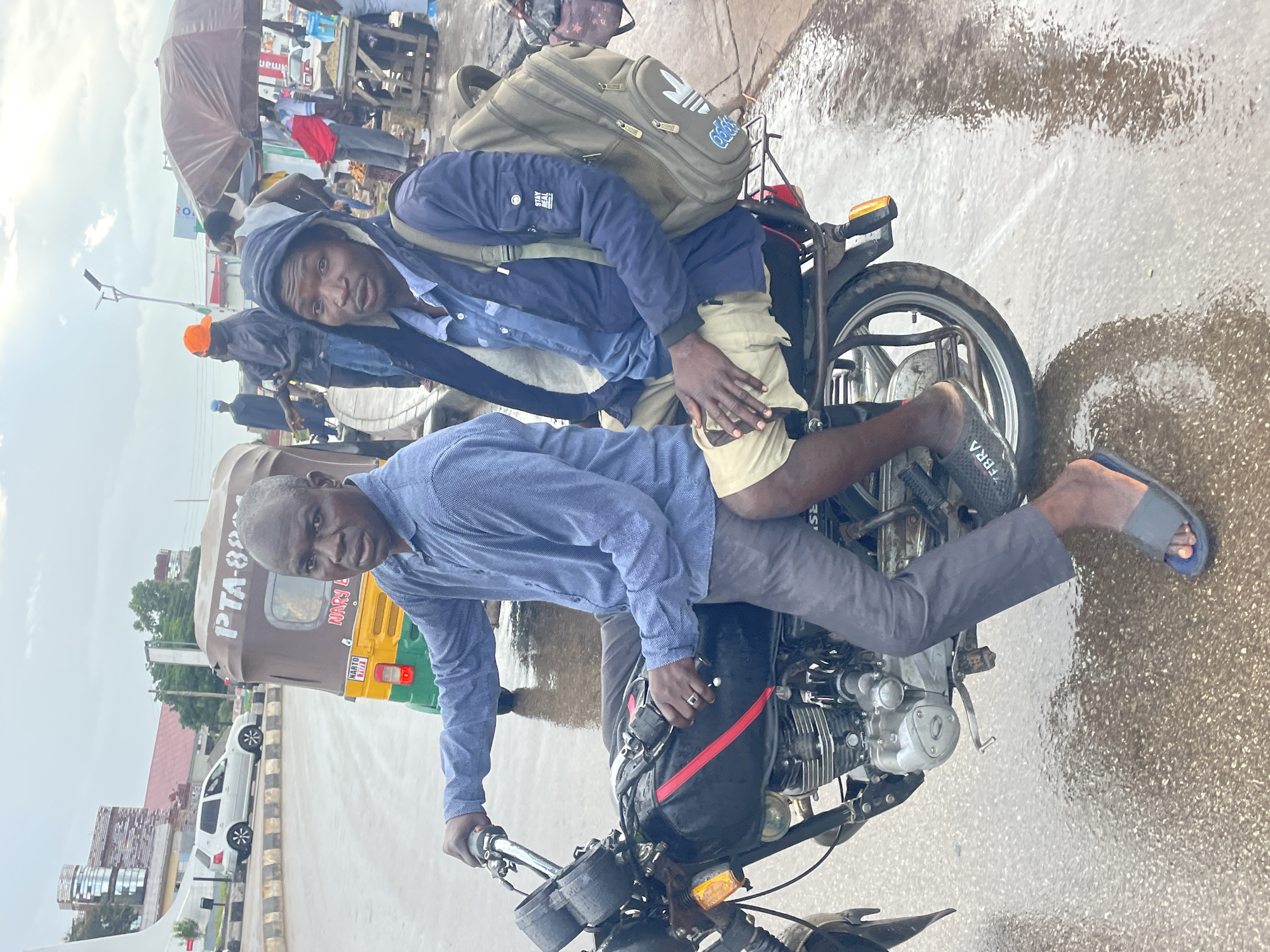 Two men are seated on a black motorcycle. The man in front, driving, is wearing a blue long-sleeved shirt and dark grey pants. The man behind him, the passenger, is wearing a blue hooded jacket over a light blue collared shirt and tan shorts. He has a large, olive-green backpack on. Both men are looking forward, and their faces are visible. The motorcycle is parked on a wet, paved road, suggesting it may have recently rained. In the background, there is a brown and yellow auto-rickshaw with "PTA-8891" visible on its side. Further in the background, a white SUV is driving on the road, and there are buildings and market stalls with umbrellas. The overall environment appears to be an outdoor urban setting, likely in Jos, Nigeria, given the presence of auto-rickshaws common in the region and typical market scene. The sky is overcast, contributing to the muted lighting.