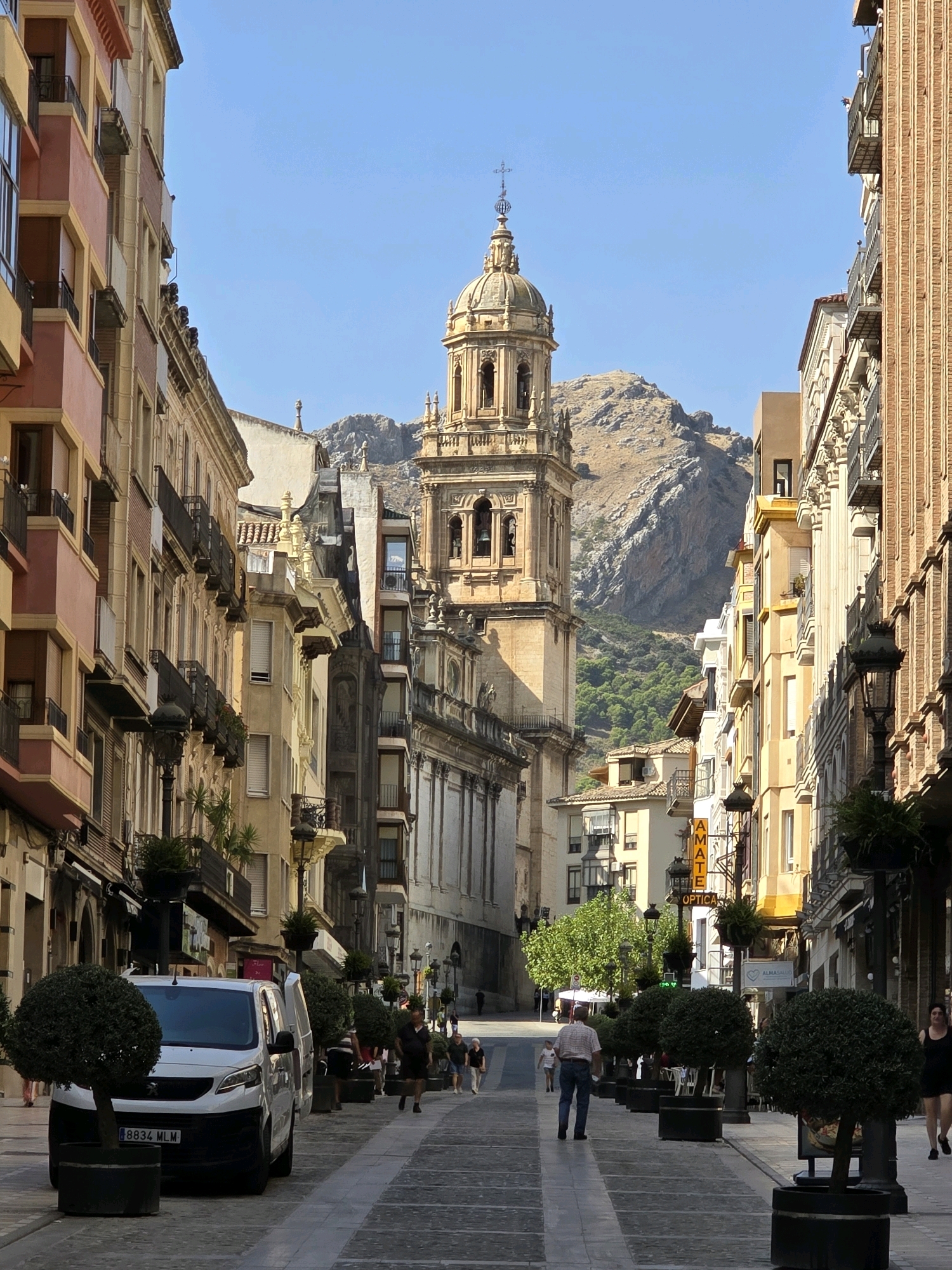 The image depicts a street in the city of Jaén, Spain, leading towards a prominent church with a dome and bell tower. The architecture of the surrounding buildings is a mix of classical and modern styles, with balconies and ornate details. The street is paved with cobblestones and lined with trees in planters, as well as lampposts. Several people are visible walking along the street. A white van is parked on the left side of the street. In the background, a rocky mountain looms over the city. The sky is clear and blue, suggesting a sunny day. The lighting indicates that the sun is high in the sky, casting shadows on the buildings and street.