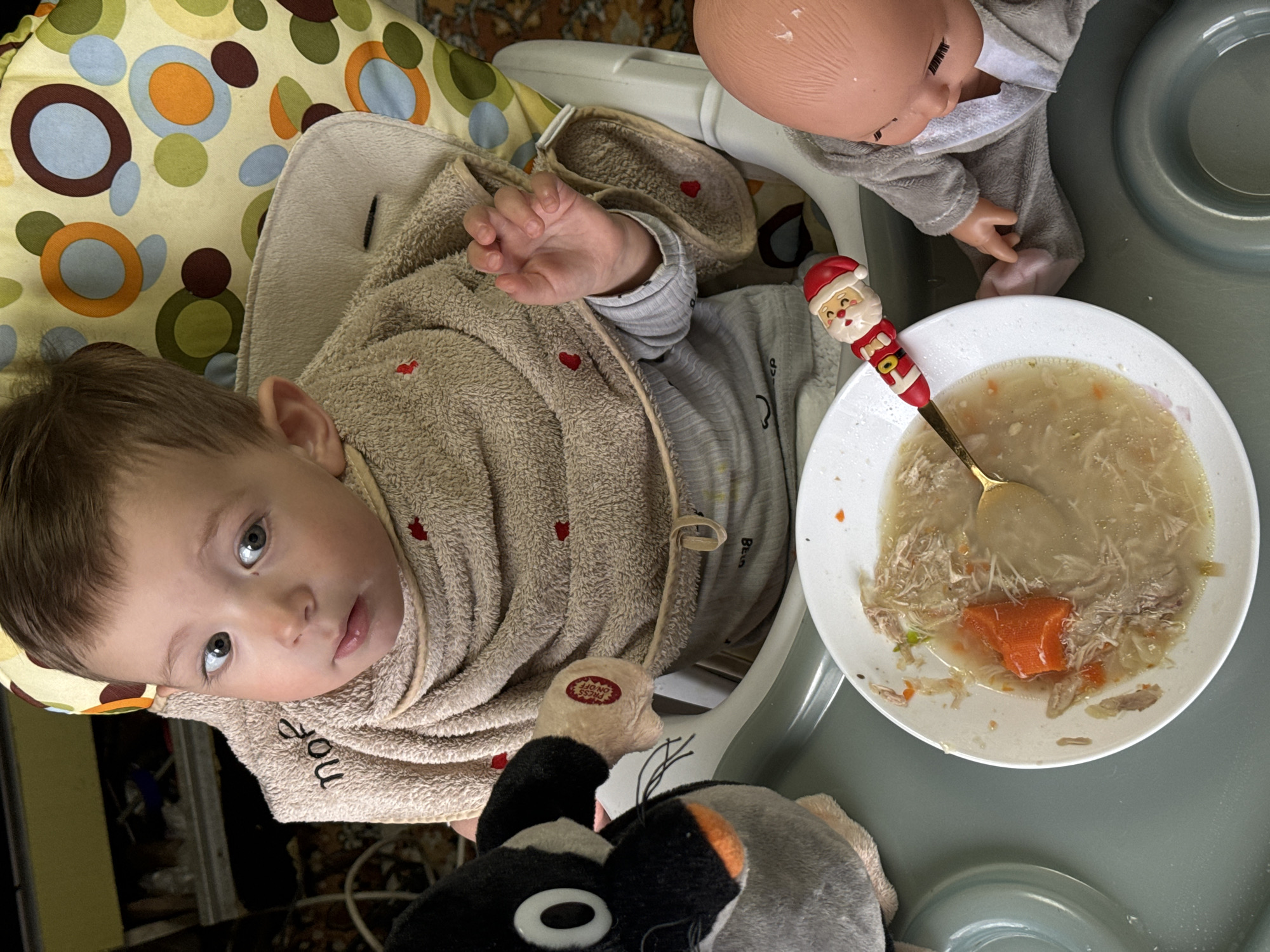 A young child with light brown hair and blue eyes sits in a high chair, looking upwards. The child is wrapped in a beige towel with small red heart designs. A plate of soup with shredded chicken and a carrot is on the tray, along with a spoon that has a Santa Claus figure on the handle. Next to the soup bowl is a doll dressed in grey. Below the child is a stuffed animal with black and white features. The high chair has a patterned fabric seat with circles of various colors. The scene appears to be indoors. Location: Unknown, Unknown.