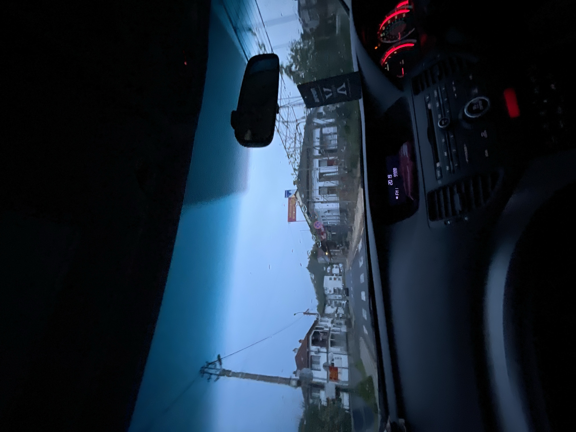 The image is taken from inside a vehicle, likely a car, looking out through the windshield. It appears to be a cloudy or rainy day. The vehicle seems to be on a street in what looks like a residential area or a small town. Based on the orientation of the photo, the vehicle is driving perpendicular to the buildings on a street in Zlatograd, Bulgaria. There are buildings, utility poles with overhead wires, and possibly some trees visible. The rearview mirror and a hanging ornament are seen on the windshield. The dashboard and part of the steering wheel are also visible. The instrument panel shows a red glow, indicating that the vehicle is on. The radio display shows "FM1" and "9900" with a timestamp of "20:19."