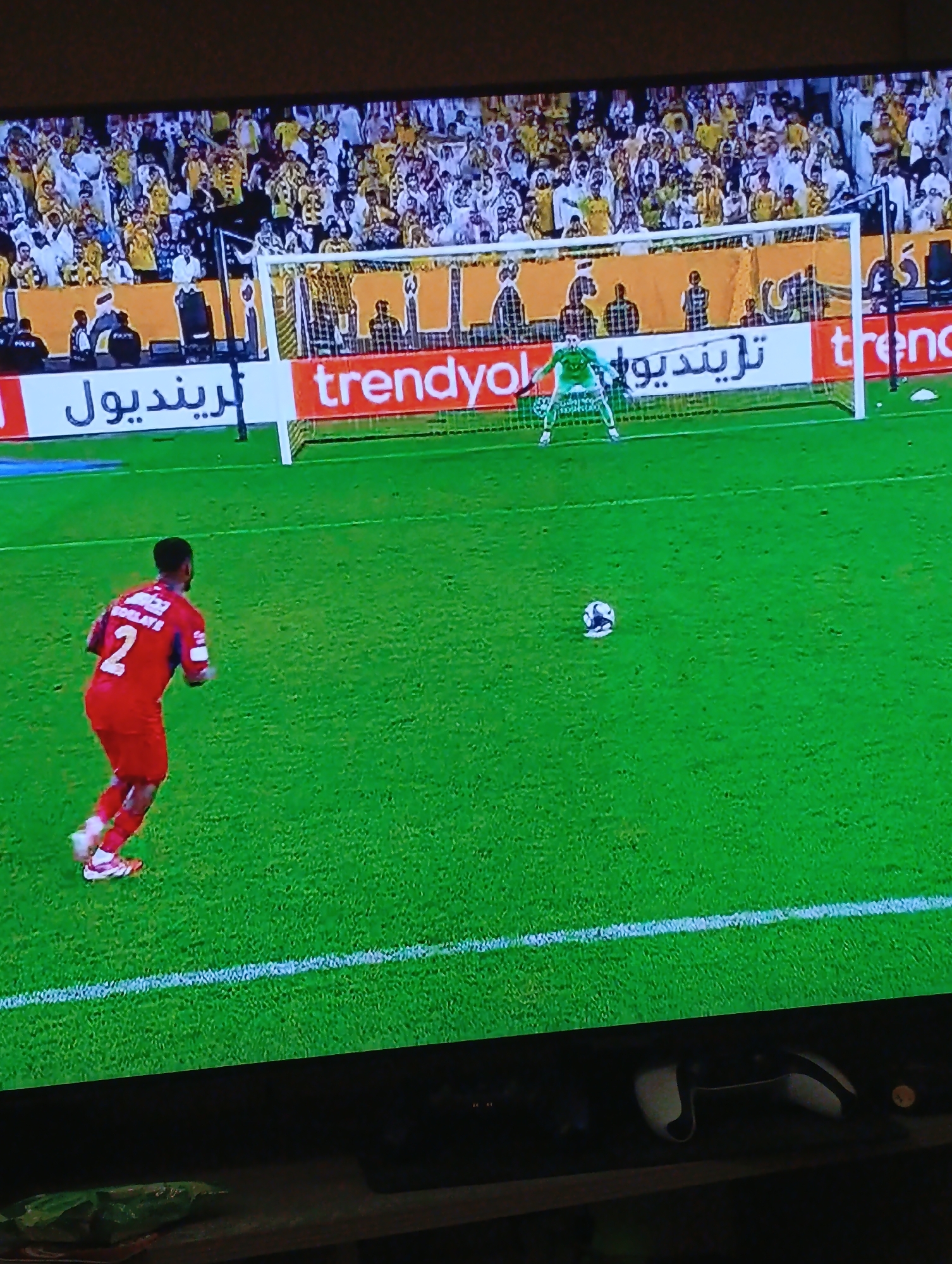 The image depicts a soccer game in progress, viewed on a television. A player wearing a red jersey with the number 2 is running towards a soccer ball in the center of the field. The goalie, wearing a green uniform, stands in front of the goal. The goal is flanked by "trendyol" advertisements in English and Arabic. A crowd is visible in the background, primarily wearing yellow and white. The field is green with white lines marking the boundaries. The location is not discernible from the image, so it is impossible to ascertain if it is in the city of Plovdiv, Bulgaria.