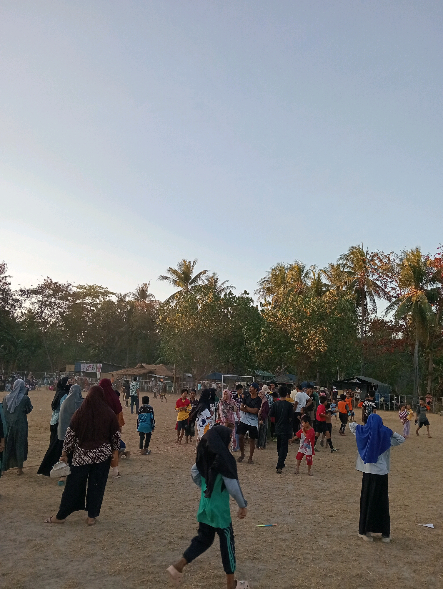 The image depicts a community gathering at an outdoor venue, likely a sports field or park, in Pagerungan Kecil, Indonesia. The scene is characterized by a dry, sandy ground and is framed by lush green trees and palm trees, suggesting a tropical or subtropical environment. The sky is a clear, pale blue, indicative of daylight, possibly late afternoon or early morning given the soft lighting.
A diverse group of people, including men, women, and children, are dispersed across the open area. Many are dressed in casual clothing, with a notable presence of women and girls wearing hijabs. Some individuals appear to be engaged in activities such as playing or conversing, while others are observing. There are several makeshift structures or stalls visible in the background, which could be temporary vendors or event booths. The overall atmosphere suggests a relaxed, communal event.