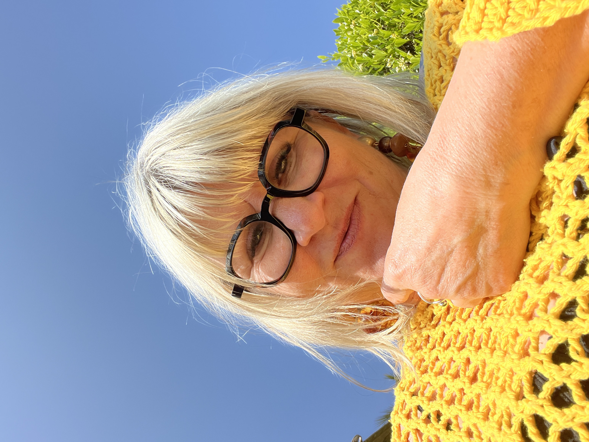 A woman with blond hair and glasses poses with her face angled downward toward the camera. She wears a yellow crocheted garment. The sky is visible behind her, along with green foliage. The location may be Grimaud, France.