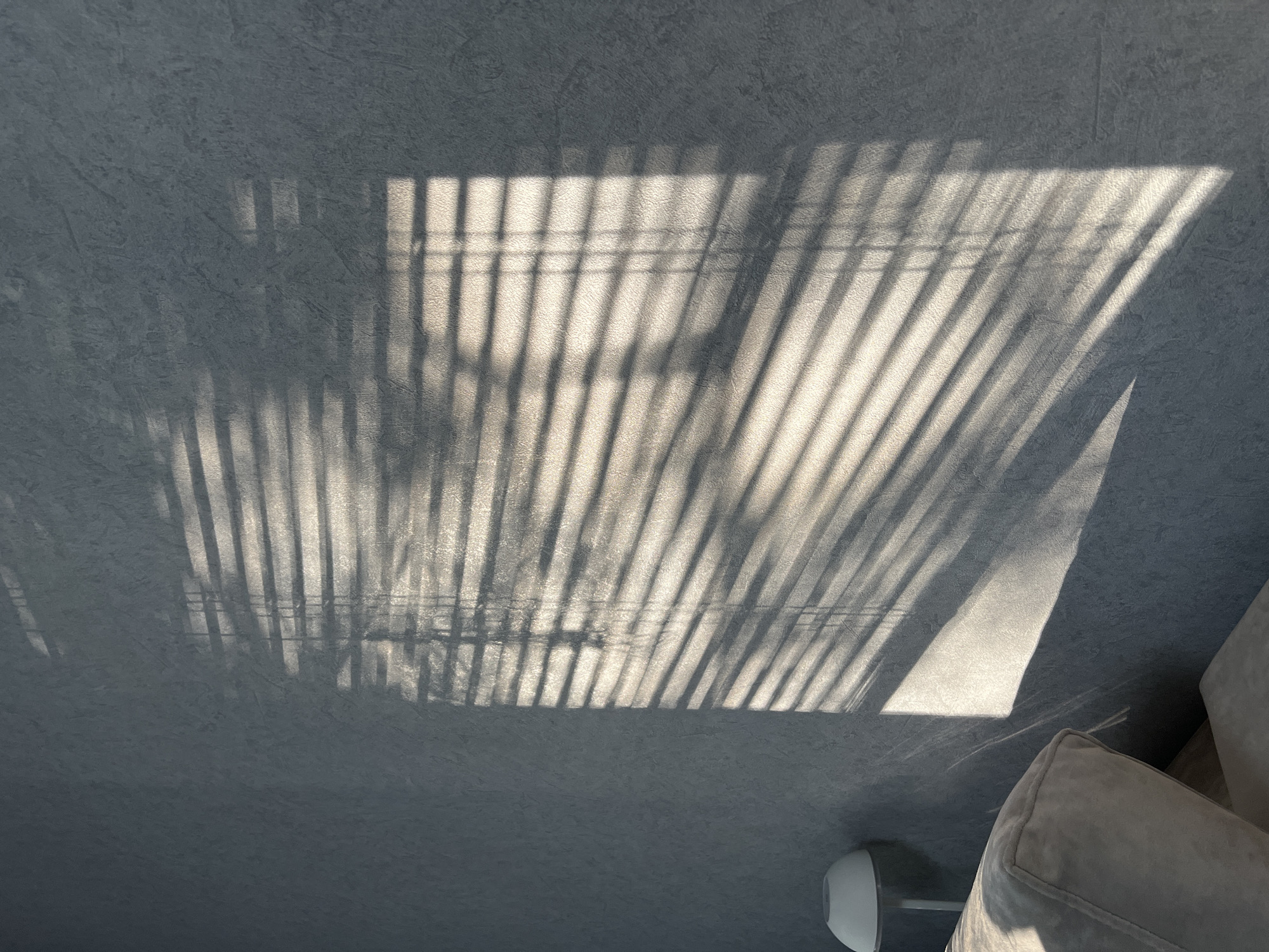 The image is a close-up shot of a textured gray wall with shadows cast by horizontal blinds. The lighting suggests it is either sunrise or sunset. In the lower left corner, a portion of a light-colored sofa and a white lamp are visible. The scene appears to be indoors, likely in a residential setting. There are no people present in the image.