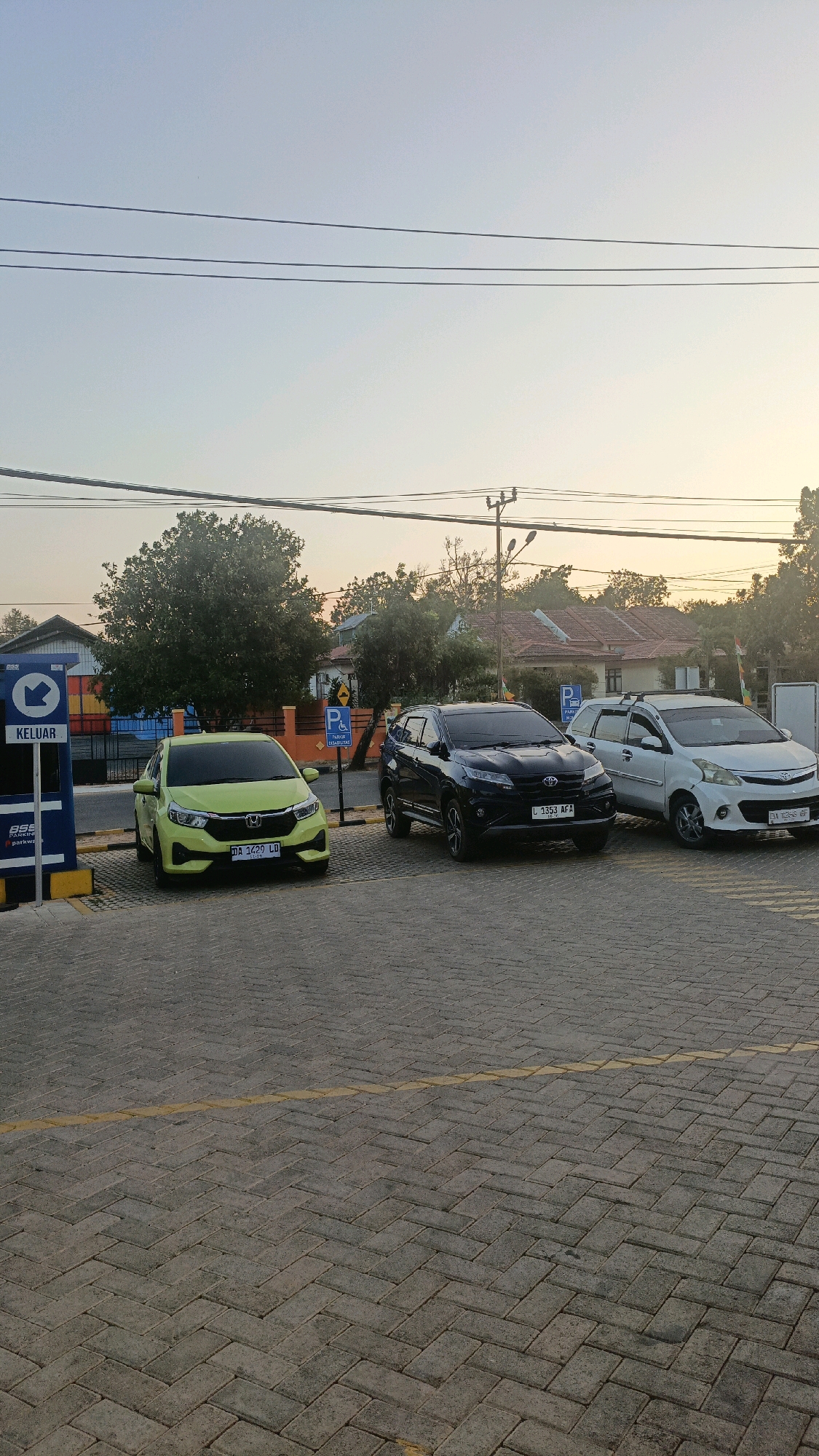 In Pelaihari, Indonesia, on a paved parking lot with a herringbone pattern and a yellow dividing line, three cars are parked. The car on the left is a light green hatchback, with the license plate "DA 1429 LD". Next to it, to the right, is a black SUV with the license plate "L 1353 AFA". The car on the far right is a white minivan, with the license plate partially visible as "DA 1255 BF". A blue sign with a white arrow pointing left and the word "KELUAR" (Exit) is visible on the left side of the image, next to a blue parking meter. Further in the background, there is a row of trees, buildings with orange tile roofs, and utility poles with wires. The sky is a pale blue transitioning to yellow near the horizon, indicating either sunrise or sunset.