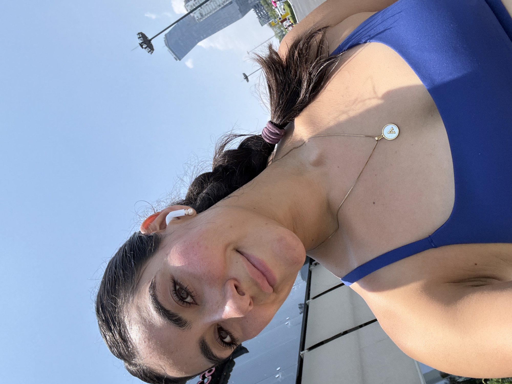 A selfie of a woman with brown hair in a braid, wearing a blue top and a necklace with a round pendant, shows her standing outdoors under a clear blue sky. She has light skin, brown eyes, and is wearing a white earbud in her right ear. In the background, there are tall buildings, one of which is notable for its unique curved shape. The location is Puebla, Mexico, identified by the architectural style of the buildings and the general urban landscape.