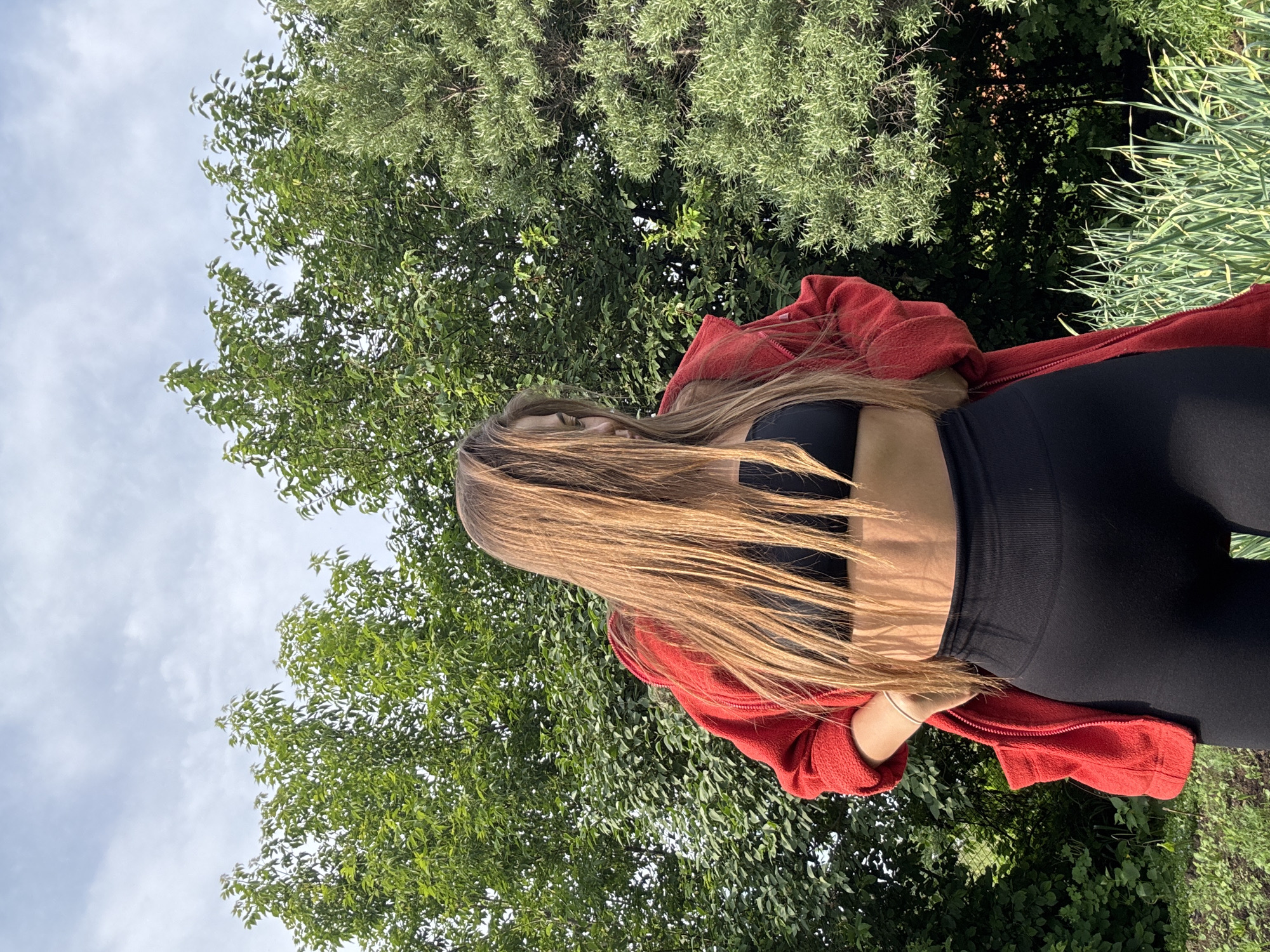 In an outdoor setting, likely a garden or yard, a young woman with long, light brown hair stands facing slightly away from the camera. Her hair is falling forward, obscuring part of her face. She is wearing a black sports bra and black leggings. Over her shoulders, she has a red, textured jacket or cardigan draped open, revealing her midriff. Her hands are placed on her hips. Behind her, a dense green backdrop of trees and foliage is visible under a partially cloudy sky. To the right, there is a larger evergreen tree with lighter green foliage. In the foreground, green plants, possibly chives or onions, are growing. The lighting suggests it is daytime, with sunlight casting shadows. The location is identified as Sulmash, Russia.