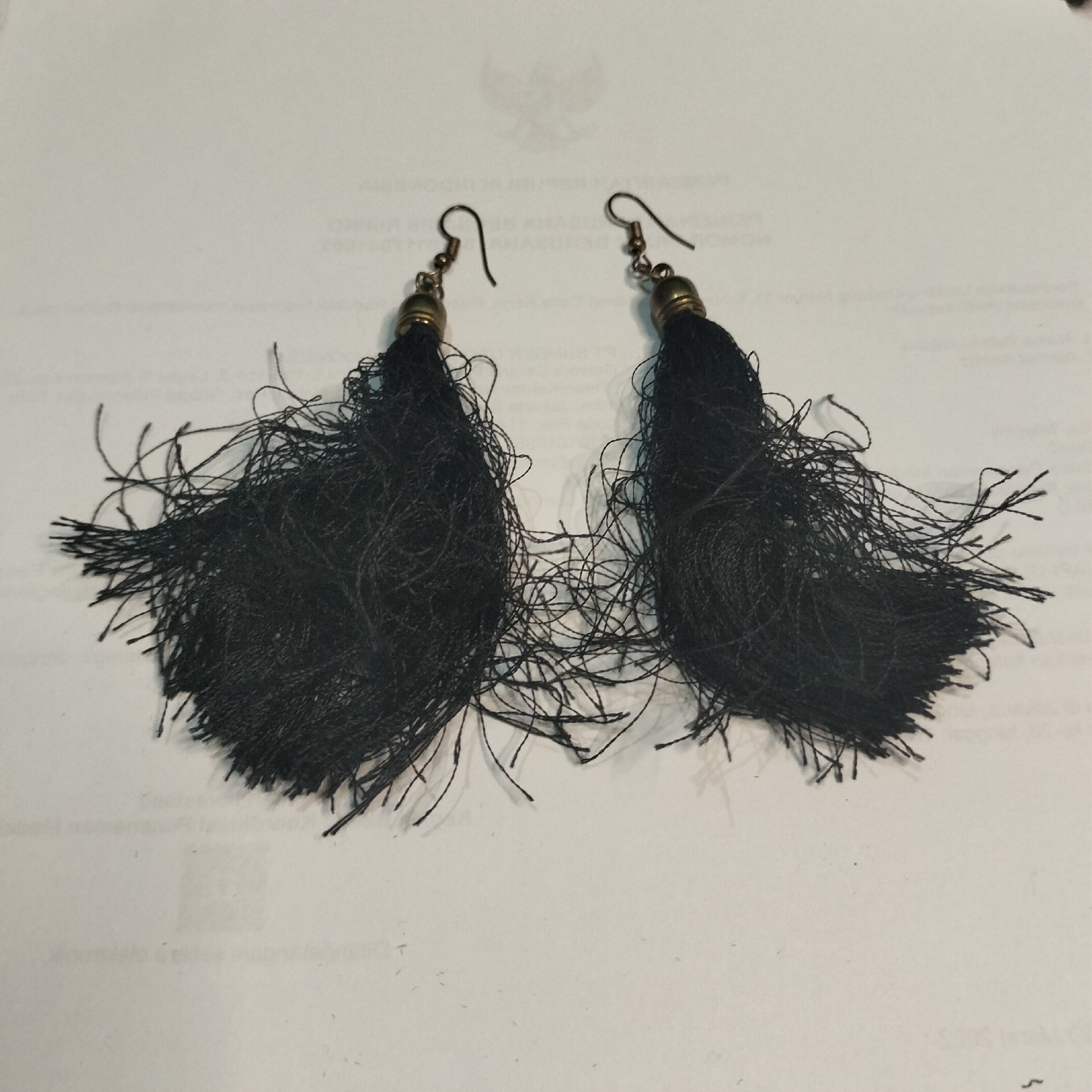 The image displays a pair of black tassel earrings resting on a white surface, possibly paper with faint text. The earrings are attached to metallic hooks, with a gold-colored cap at the top of each tassel. The tassels themselves are composed of many thin, dark threads that are somewhat frayed and spread out, giving them a wispy appearance. The background is evenly lit, showing no discernible details about the environment or location. There are no people or any specific objects that would indicate a location in Tangerang, Indonesia. The image focuses solely on the jewelry.