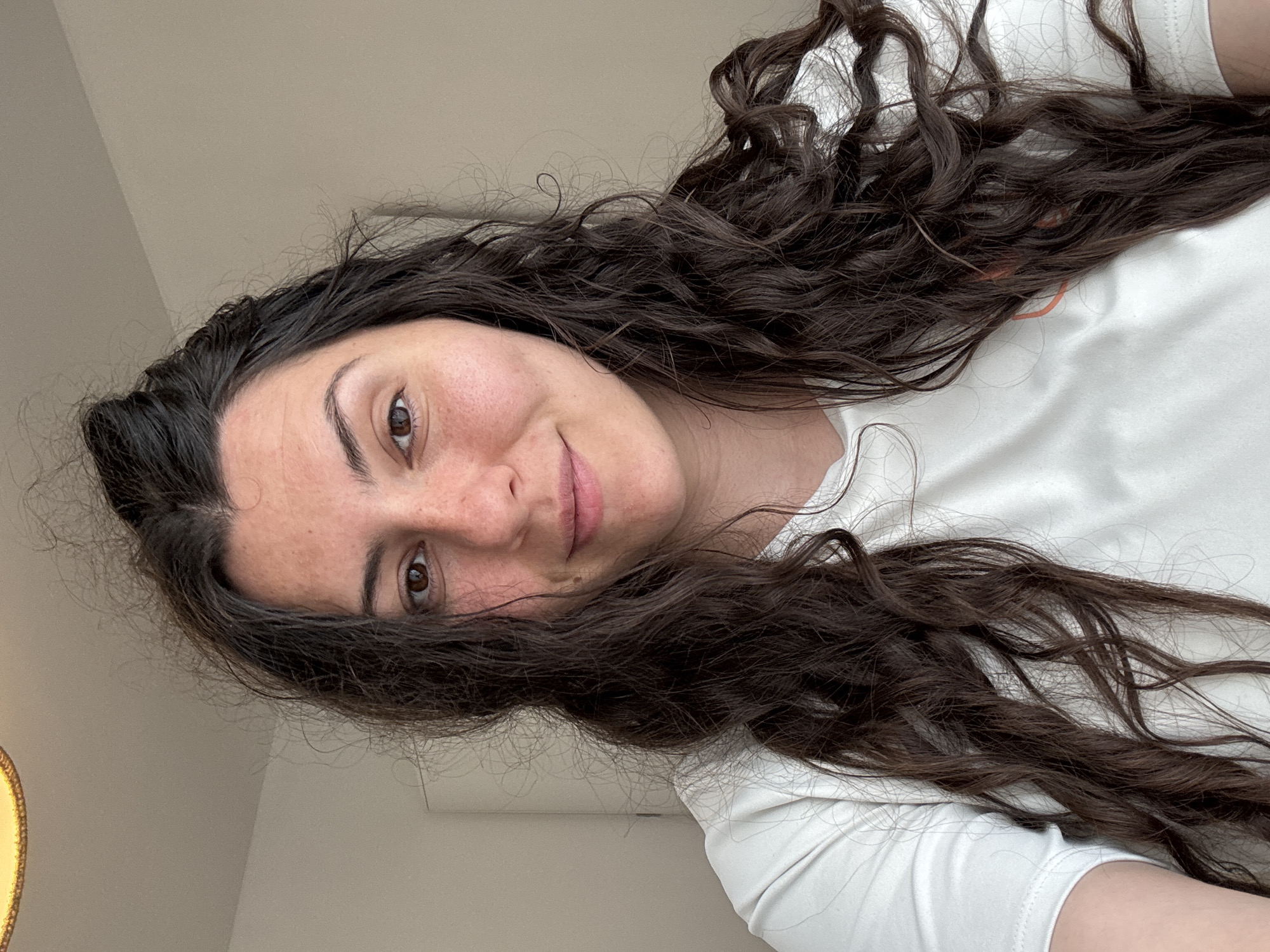 A woman with long, dark, curly hair and fair skin is shown in a selfie. She has brown eyes and freckles on her face. She is wearing a white t-shirt. The background appears to be a neutral-colored wall, possibly indoors. A lamp is visible in the lower left. The photograph is slightly tilted. The location is Unknown, Unknown.