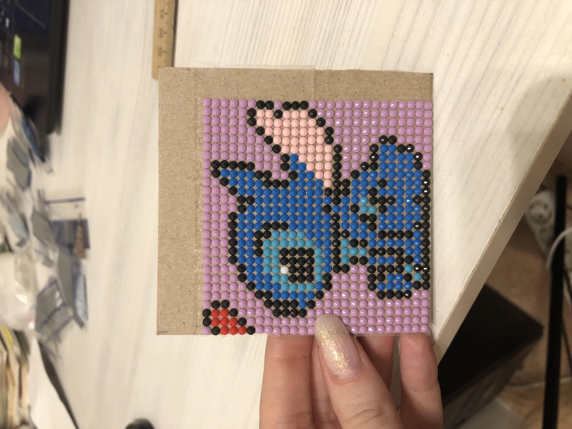 The image shows a craft project, specifically a square picture made of small, colored beads. The subject of the picture is a stylized representation of the character Stitch from the Disney movie "Lilo & Stitch." The background of the Stitch image is lavender, while Stitch himself is primarily blue and black, with a touch of pink on his head and a red detail near the bottom. The bead art is on a brown cardboard square. A hand with long, sparkly fingernails is holding a corner of the cardboard square. The background is a light-colored surface with a hint of a desk and other items. There is no indication of the location being Noviy, Russia.