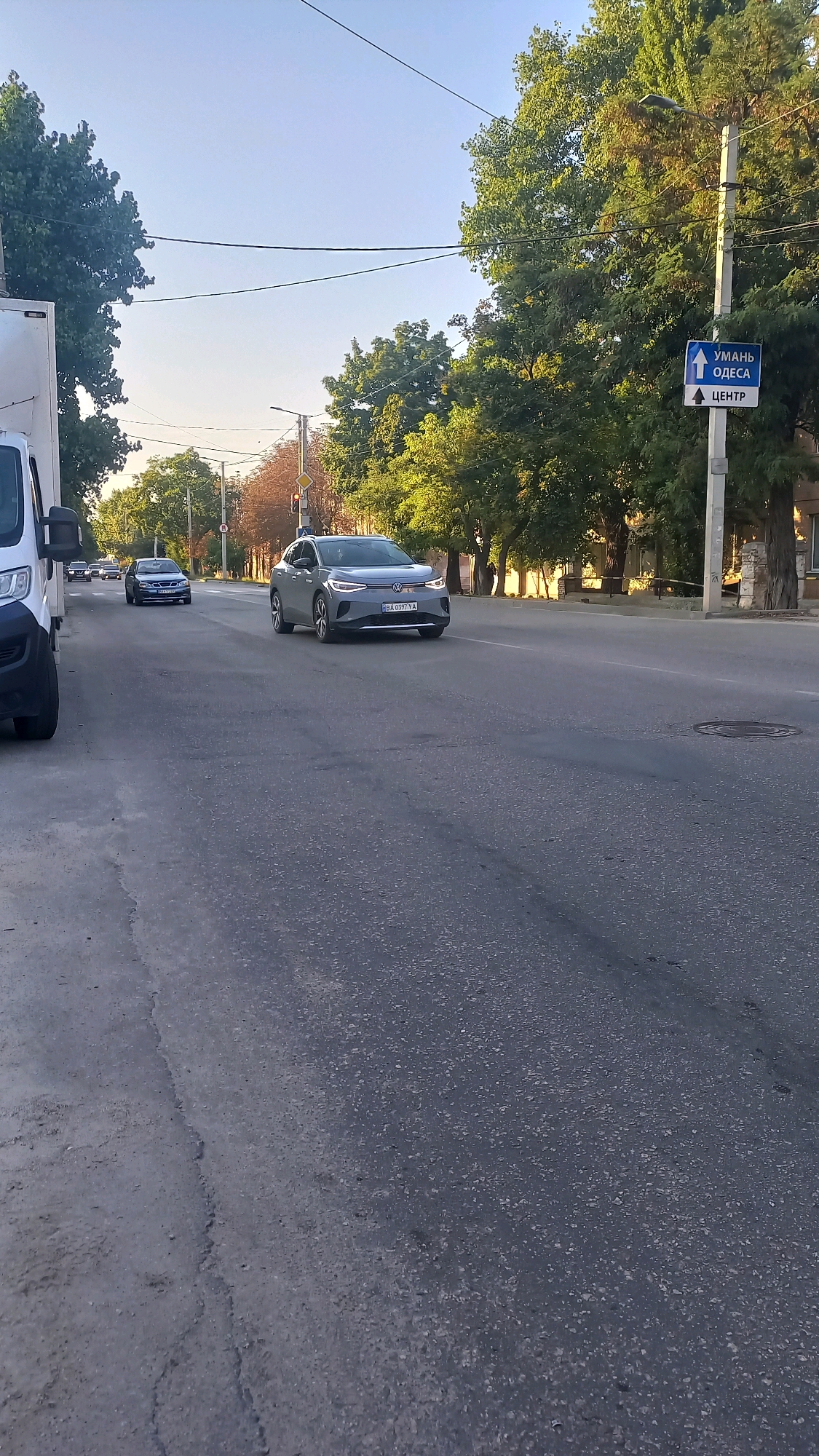 The image shows a street scene in Kropyvnytskyi, Ukraine, with several cars on a paved road under a clear blue sky. The road has visible lane markings and appears to be a multi-lane thoroughfare. On the right side of the road, a street sign indicates directions to "UMAN," "ODESA," and "CENTER" with arrows. Green trees line the right side of the street, and some buildings are visible in the background. A grey electric car, possibly a Volkswagen ID.4, is the most prominent vehicle in the foreground, driving in the right lane. Other cars are visible further down the road, moving in the same direction. On the left side of the image, a white van is partially visible, parked on the side of the road. The overall impression is that of a typical urban street scene during daylight.
