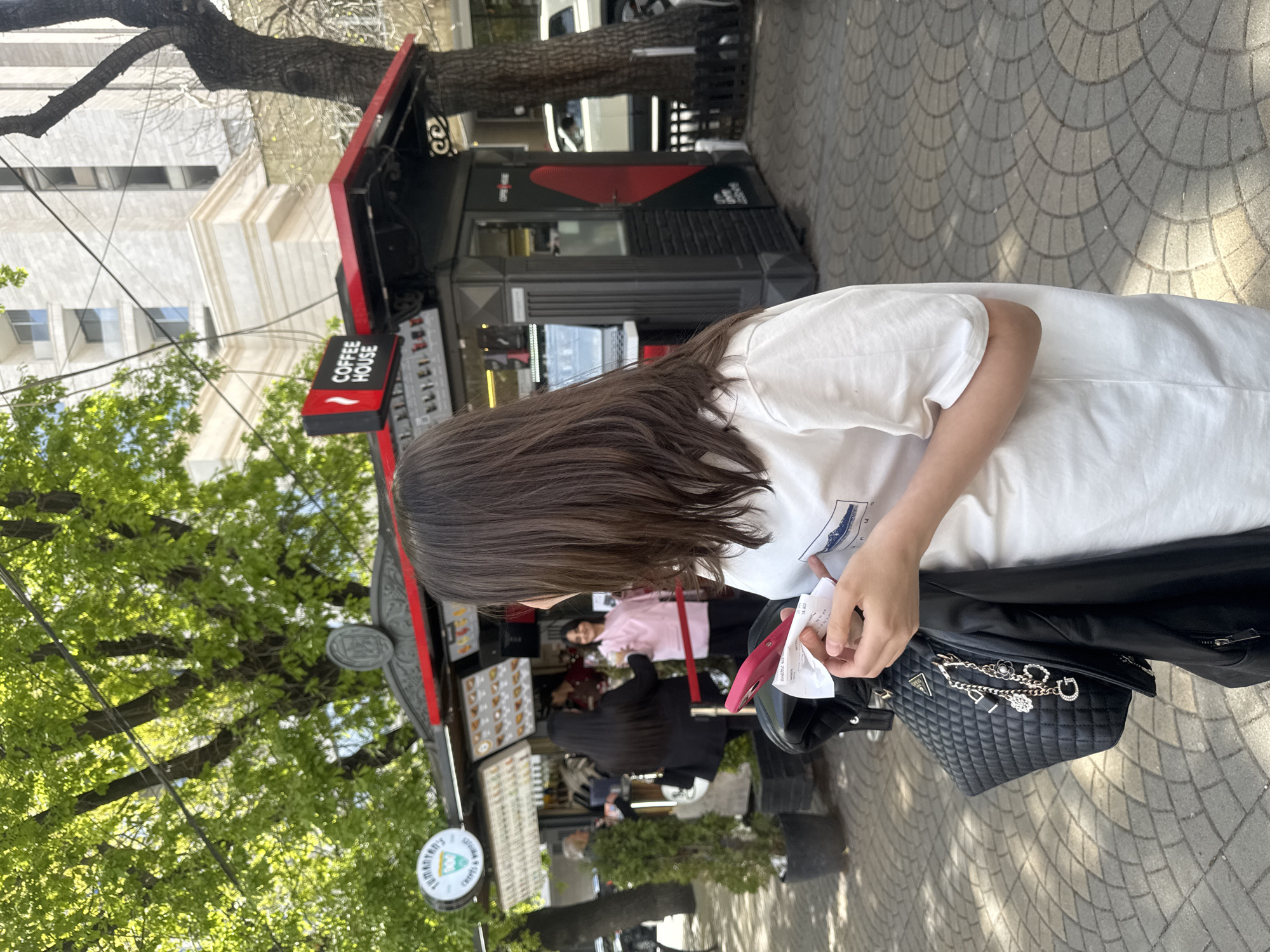 This image, taken in Yerevan, Armenia, depicts a vibrant outdoor urban scene on a bright, sunny day, likely during late morning or afternoon. The foreground is dominated by a person seen from behind, with long dark hair, wearing a white t-shirt. They are holding a pink smartphone and a white paper, possibly a receipt, while a black quilted shoulder bag with chain straps and charms (including a visible 'G' indicating a Guess brand) rests on their arm, along with a black leather jacket.
The central feature of the scene is a bustling street-side coffee kiosk, primarily black with red accents. A prominent sign on the kiosk reads "COFFEE HOUSE" in white letters with a white wavy logo. Below its awning, a circular sign advertises "TUMANYAN'S / CREPES & COFFEE" with a green logo, indicating the kiosk offers both beverages and food.
Several other individuals are visible around the coffee stand; one person in a pink top and another with long dark hair are seen near the serving area, suggesting customer interaction or service. The ground is a patterned paved sidewalk. Lush green trees provide natural elements and shade, while a light-colored, multi-story building with windows stands in the background. Overhead utility wires are also present. The overall activity revolves around people gathering at the kiosk for coffee and crepes in a casual city setting.