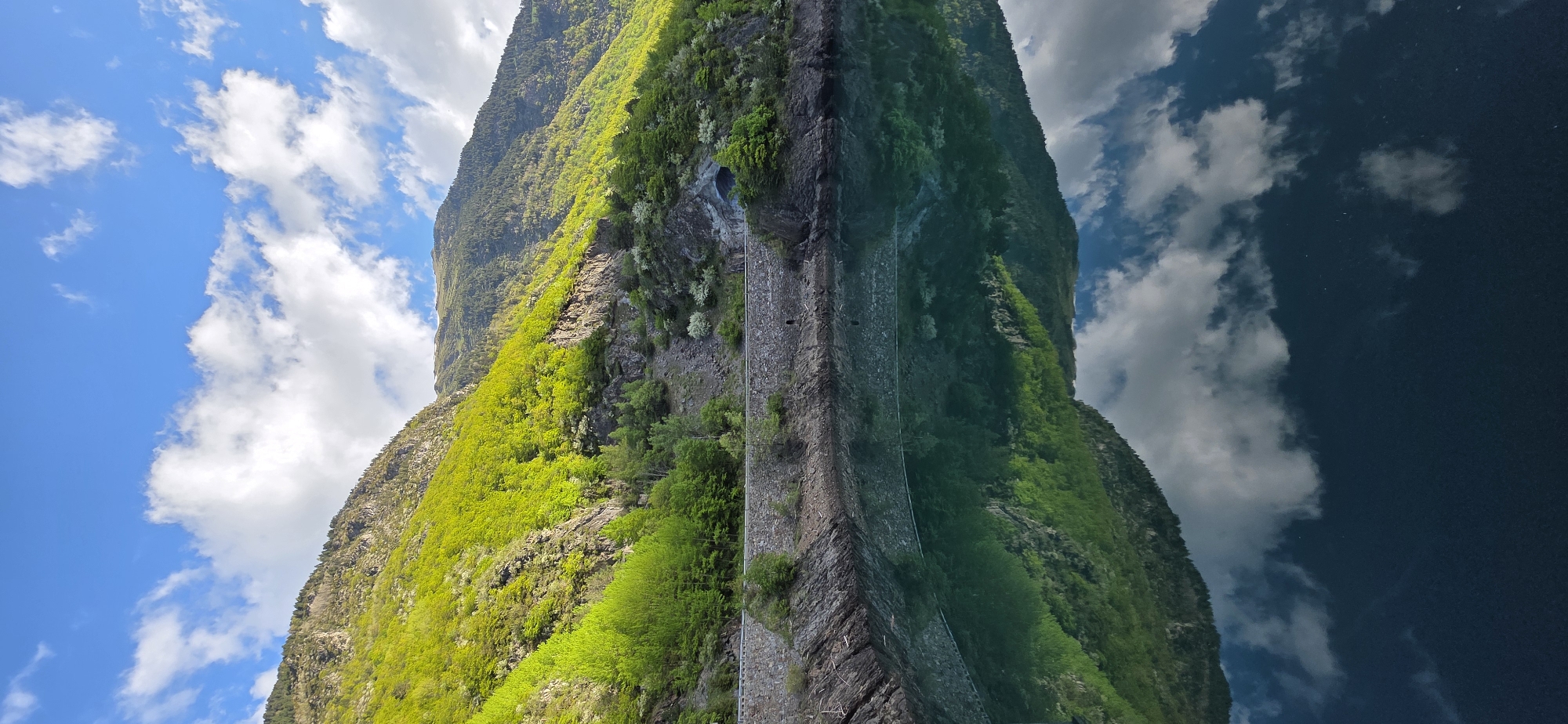 The image depicts a mountain reflected in a body of water, likely a lake or fjord. The mountain is covered in green vegetation, and a sheer rock face runs down its center. The reflection in the water mirrors the mountain, sky, and clouds above, creating a symmetrical composition. The sky is blue with white clouds. The reflection of the sky is darker in the water. The location is in the city of Benasque, Spain.