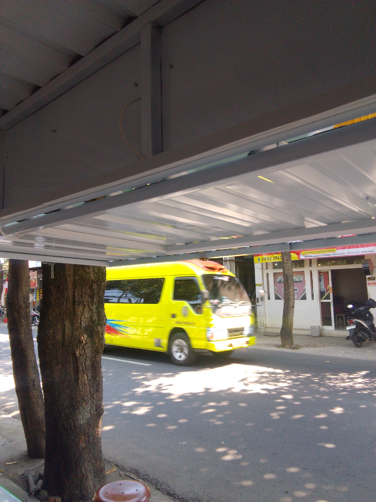 The image captures a street scene in Trenggalek, Indonesia. A bright yellow minibus, likely a public transportation vehicle, is in motion on the asphalt road, its movement suggested by the slight blur. The bus features colorful decals along its side. In the foreground, the textured bark of two trees stands prominently, with dappled sunlight filtering through their leaves, casting shadows on the road.
Behind the trees, a shop or business is visible with its entrance. Signage on the building indicates it offers printing services, including banners, stickers, and ID cards. A phone number, "0853 3513," is also displayed. A black motorcycle is parked near the shop. The overall atmosphere suggests a daytime scene, with good weather, as indicated by the clear shadows and bright lighting. The setting appears to be an urban or semi-urban area, given the presence of businesses and paved roads.