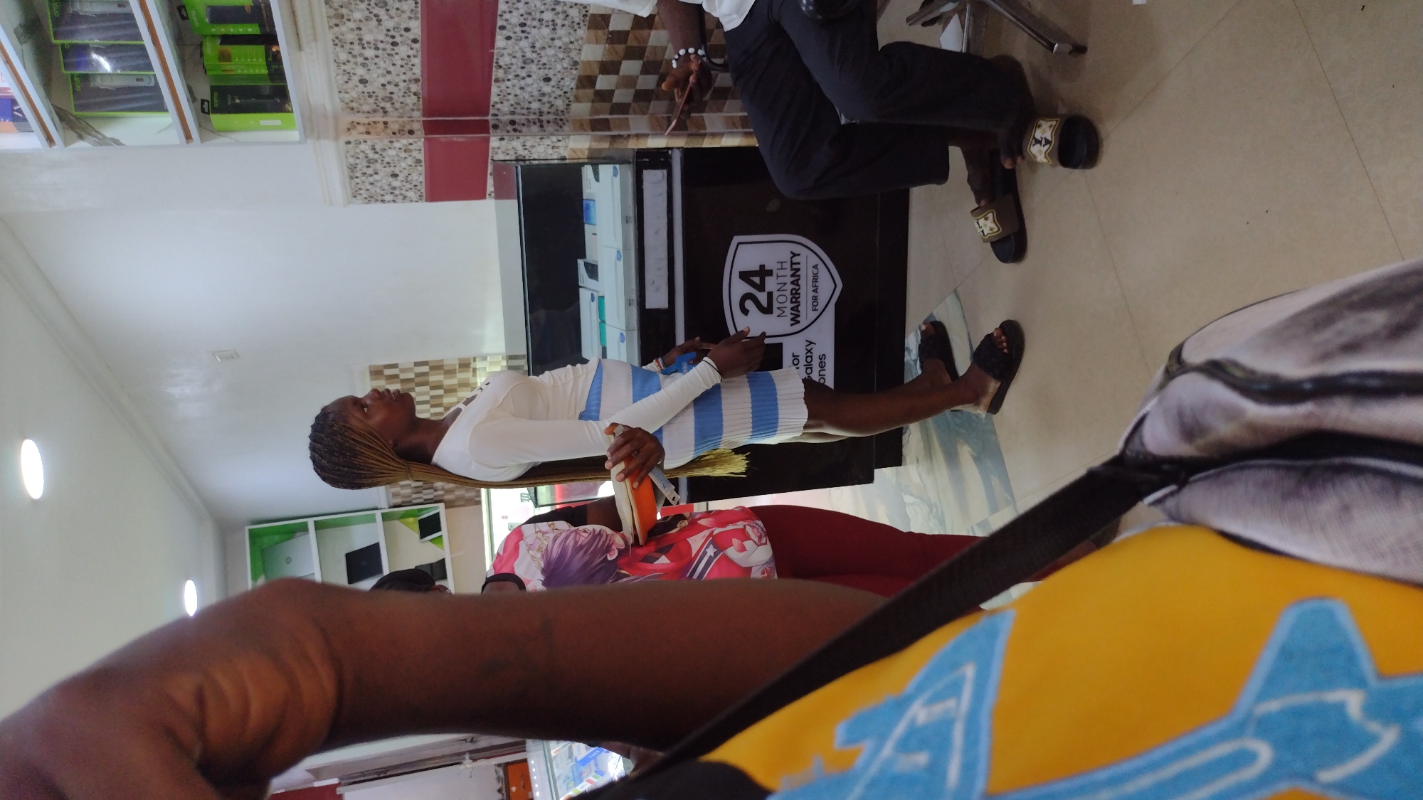 The image shows the interior of a store, possibly a mobile phone shop, in Jos, Nigeria. A woman in a white top and striped dress stands in front of a display cabinet that advertises "24 Month Warranty for Africa". Another person wearing black pants is seated nearby. Other customers are also in the frame. The foreground is partially obscured by the shoulder and bag strap of the person taking the picture. The shop has white walls and tiled accents. Shelves with packaged items are visible in the background.