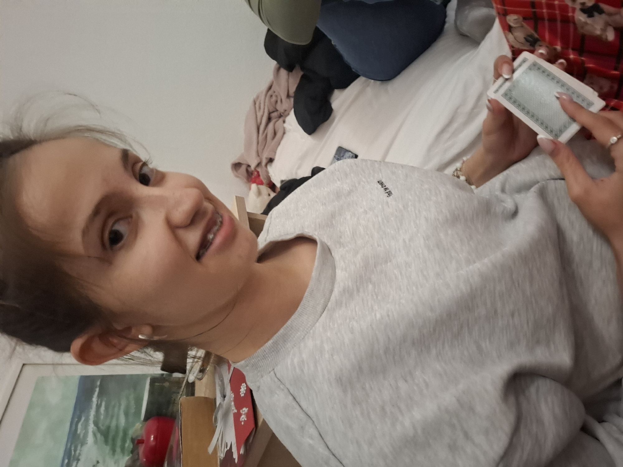A young woman with brown hair and braces is holding playing cards. She is wearing a light grey sweater. The scene appears to be indoors, on a bed. The location is likely in Panagyurishte, Bulgaria, but that cannot be confirmed from the image. There is a painting or photograph visible in the lower left corner.