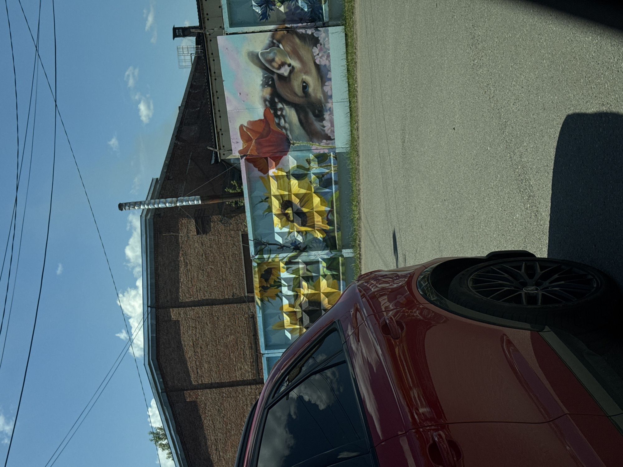 The image shows a red SUV parked on an asphalt road in Chaikovsky, Russia. In the background, there is a brick building and a mural on a fence depicting sunflowers and a deer. The sky is blue with scattered clouds. There are no people visible in the image.