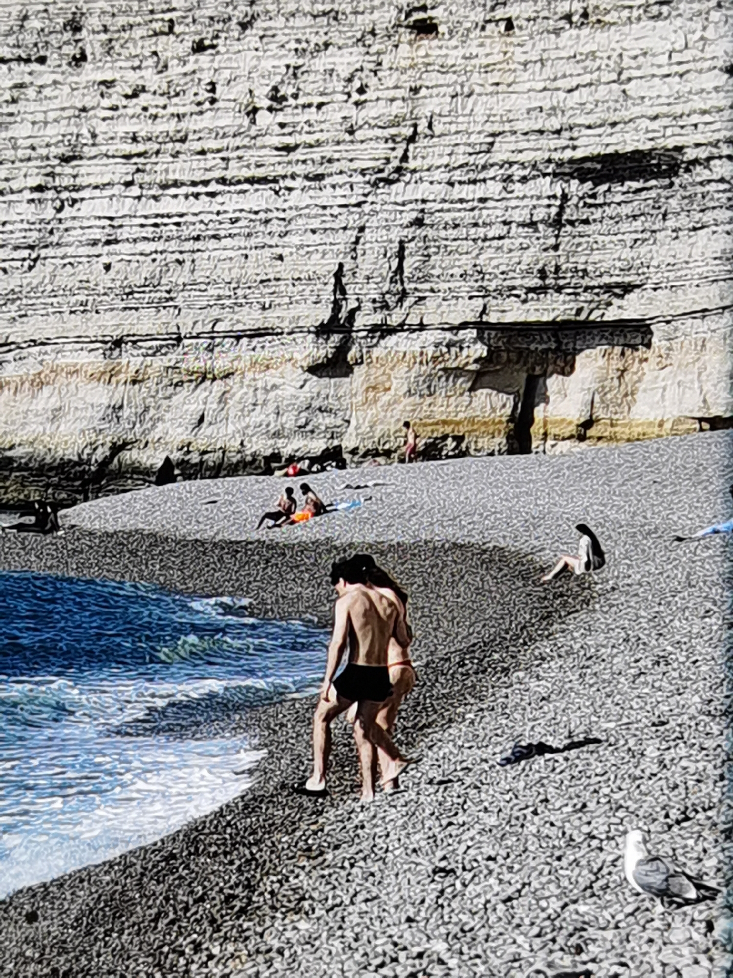 The image depicts a rocky beach in Étretat, France, with a dramatic cliff face in the background. The cliff is striated with horizontal layers of rock, appearing weathered and ancient. Sunlight casts shadows across the cliff, highlighting its rugged texture.
Several individuals are visible on the beach, engaged in various activities. In the foreground, a couple stands close together, facing away from the viewer. The male is wearing black swim trunks, and the female is wearing a red bikini. They appear to be walking or standing on the pebbly shore.
Further back on the beach, other figures are scattered. Some are seated or reclining on towels, while others are in motion. One individual, dressed in a white garment, is sitting on the pebbles. A person in black swimwear is lying down on the left side of the frame. In the middle ground, two people are seated next to each other, one wearing orange shorts.
The sea is visible on the left side of the image, with gentle waves lapping against the shore. The water is a deep blue, contrasting with the grey pebbles of the beach. A seagull stands on the pebbles in the lower right corner of the image, facing right. The overall scene conveys a relaxed atmosphere of a day at the beach.