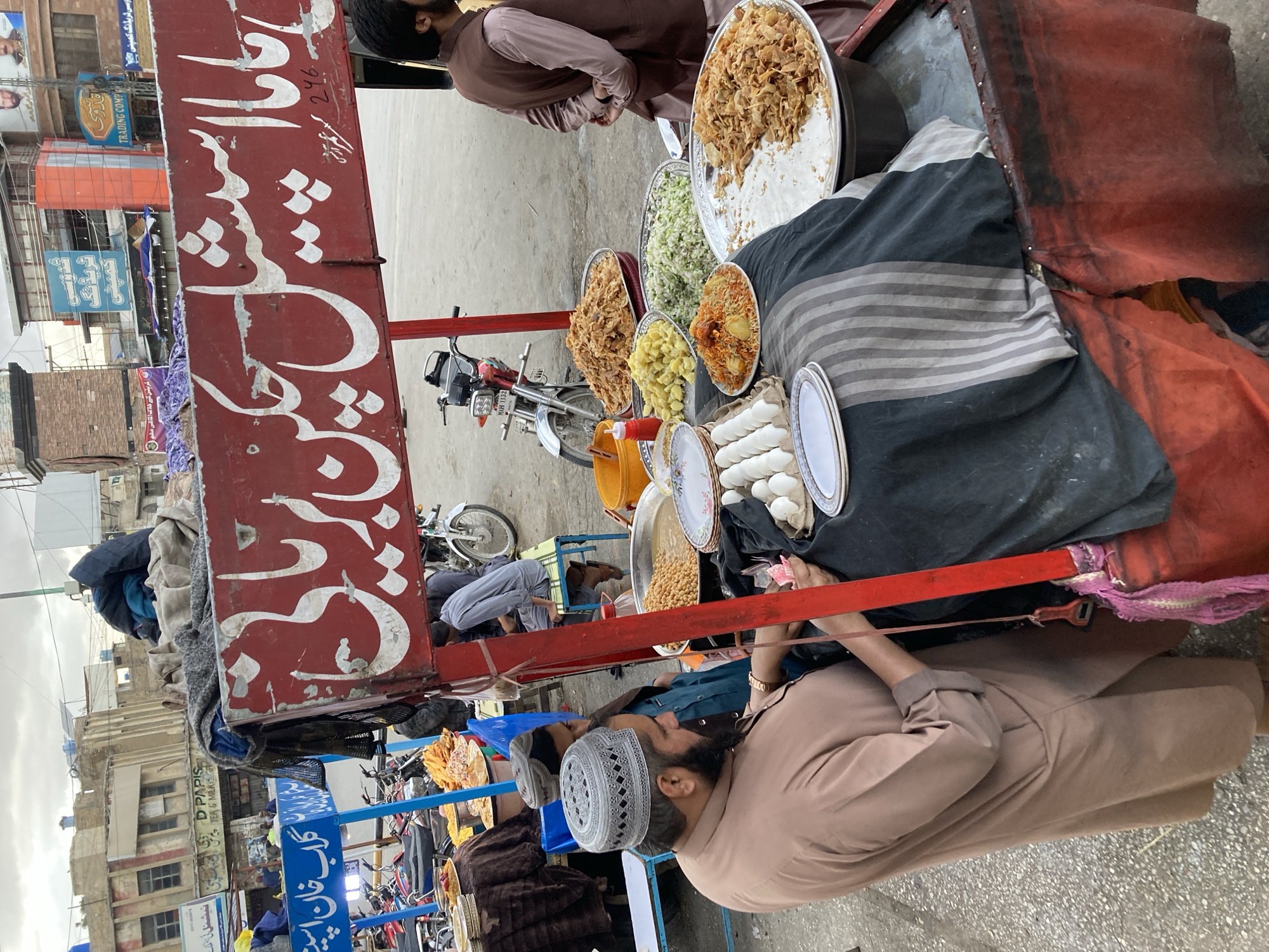This image depicts an active street food vendor scene in Quetta City Tehsil, Pakistan.
In the foreground, a red-framed food cart is positioned on a paved street. A large red sign with white Urdu script, possibly reading "Baba Special Chicken Biryani," is mounted atop the cart. The cart's counter is covered with a black and grey striped cloth, displaying various food items. These include a large metal bowl containing chickpeas in gravy, a plate of diced boiled potatoes, a plate of shredded green cabbage, a plate of biryani rice with chicken/potatoes, two large metal trays filled with fried crisp items (possibly papad or similar snacks), and a carton containing approximately eighteen white boiled eggs. A stack of white plates with a blue floral pattern and a red squeeze bottle are also present on the counter. An orange plastic bucket is visible to the right of the cart.
An adult male, presumably the vendor, stands on the left side of the cart, wearing a light brown shalwar kameez and a white patterned kufi cap. He has a dark beard and is looking down, appearing to handle small paper items, possibly currency, with his right hand. A gold-colored wristwatch is on his left wrist. A young boy in a green shirt and a dark cap stands partially obscured behind the vendor. To the right of the cart, another adult male with a beard, dressed in a brown shalwar kameez and a brown waistcoat, stands with his back to the camera, his hands clasped behind him.
In the mid-ground, several motorcycles are parked along the street, including a red motorcycle with license plate "MAH 1132". Another blue-framed food stall with Urdu script ("گلاب خان اسپی" - possibly "Gulab Khan Special") is visible to the left. Several individuals are seated or standing near this stall.
The background features multi-story urban buildings, some appearing weathered, with various commercial signs in Urdu and English, such as "D PAPIS TEA & MILK" and "TRADING COMPANY." The sky is overcast, suggesting cloudy weather. The ground surface is paved, likely asphalt or concrete.