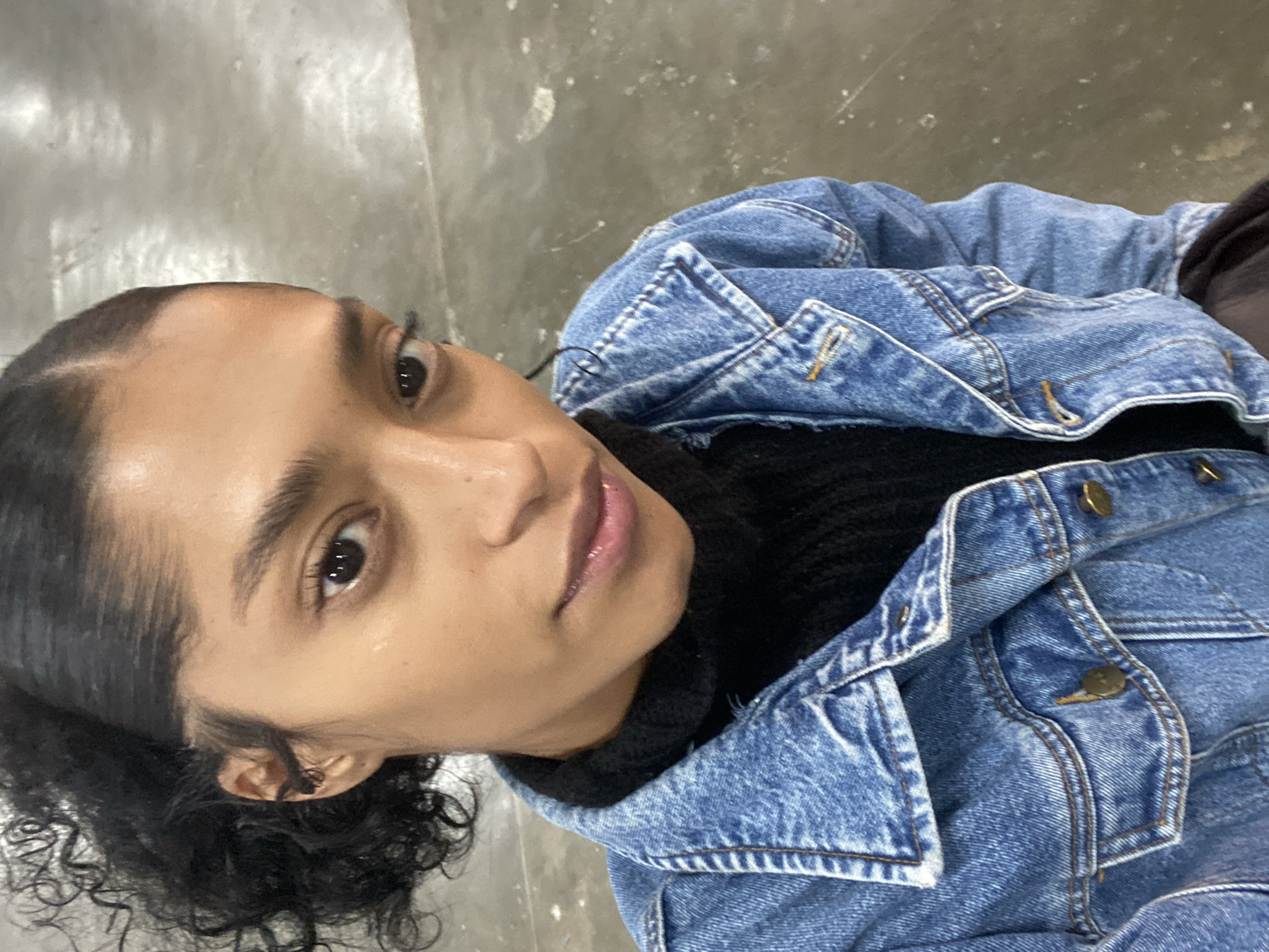 A close-up, slightly angled portrait shows a person with dark curly hair pulled back, wearing a black turtleneck sweater and a blue denim jacket. The person is fair-skinned, with dark eyes and full lips. The background appears to be a gray concrete surface, potentially a floor. The location is unknown and cannot be determined from the image.