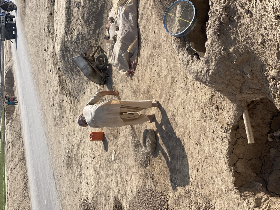 The image depicts an outdoor, arid scene in Kandahar, Afghanistan, characterized by manual labor and a roadside environment under bright sunlight. A barefoot individual, wearing light-colored traditional clothing and a head covering, is positioned centrally, actively sifting small aggregates using a flat tool and a round, shallow sieve. To their left, an orange plastic jerrycan rests on the dry, cracked earth. To their right, an overturned wheelbarrow is visible near a light-colored tarp covering a mound of material, with smaller objects placed on top. In the immediate foreground, a deep, earthen pit with fractured walls contains a horizontally placed wooden pipe. Adjacent to this pit, a large, blue-rimmed mesh sieve covers an opening within a raised, irregular earthen structure. The ground surface consists of dry, compacted, cracked soil and scattered small stones. In the background, a paved two-lane road stretches horizontally. Three children are visible standing near the far side of this road, while a light-colored truck and a motorcycle carrying two individuals are traveling further down the road. Distant earthen banks and faint green vegetation are present on the horizon. The scene is brightly illuminated, casting prominent shadows.