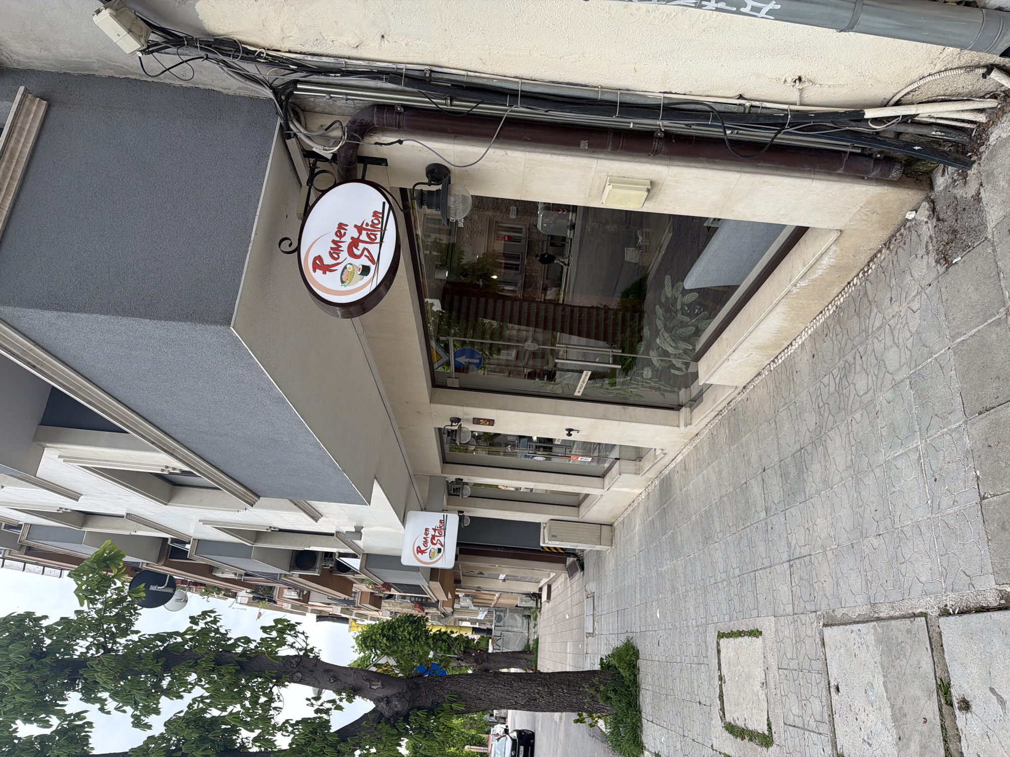 The image shows a street in Varna, Bulgaria, featuring "Ramen Station", a restaurant. The restaurant's sign is visible. The street is paved and has a tree planted on the side. There is a building with a grey overhang above the restaurant. Cables and pipes are visible along the building facade. The reflection in the shopfront shows the street scene on the other side.