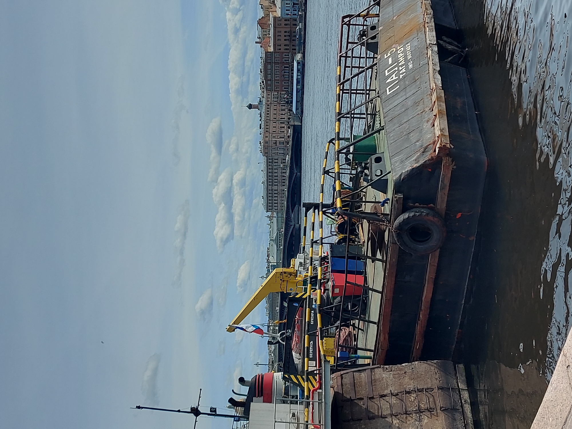 The image shows a side view of a vessel, "PAP-5" "TAGANROG", moored alongside a concrete embankment. The water level is at the base of the wall. The buildings of Saint Petersburg are visible in the background, including structures with domes, suggesting the general location is Saint Petersburg, Russia. A yellow crane is mounted on the vessel. The Russian flag is visible.