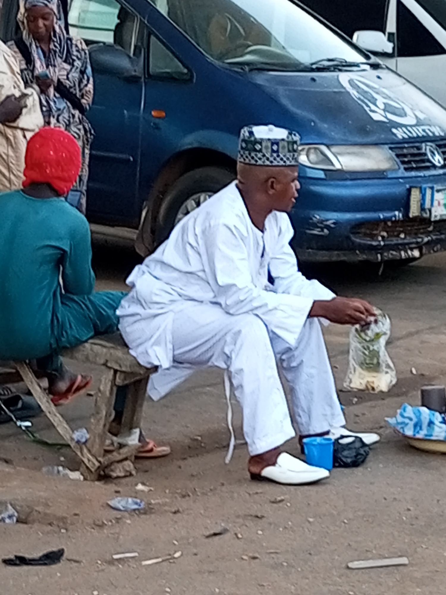 In Gombe, Nigeria, a man wearing a white cap and white traditional attire sits on a wooden bench beside a blue vehicle. His legs are crossed, and he is holding a small item in his right hand, which is enclosed in a plastic bag. To his left, another individual, wearing a red head covering and a green traditional outfit, sits with their back to the camera, facing the same direction as the man in white. The man in white is wearing white shoes with brown accents. The ground is covered in dirt and scattered debris, including plastic bags and cigarette butts. A blue bucket and a black bag are placed near the man in white's feet. In the background, the blue vehicle has a circular logo on its hood with the word "NORTH" written below it. The overall scene depicts a street-side setting in Gombe.