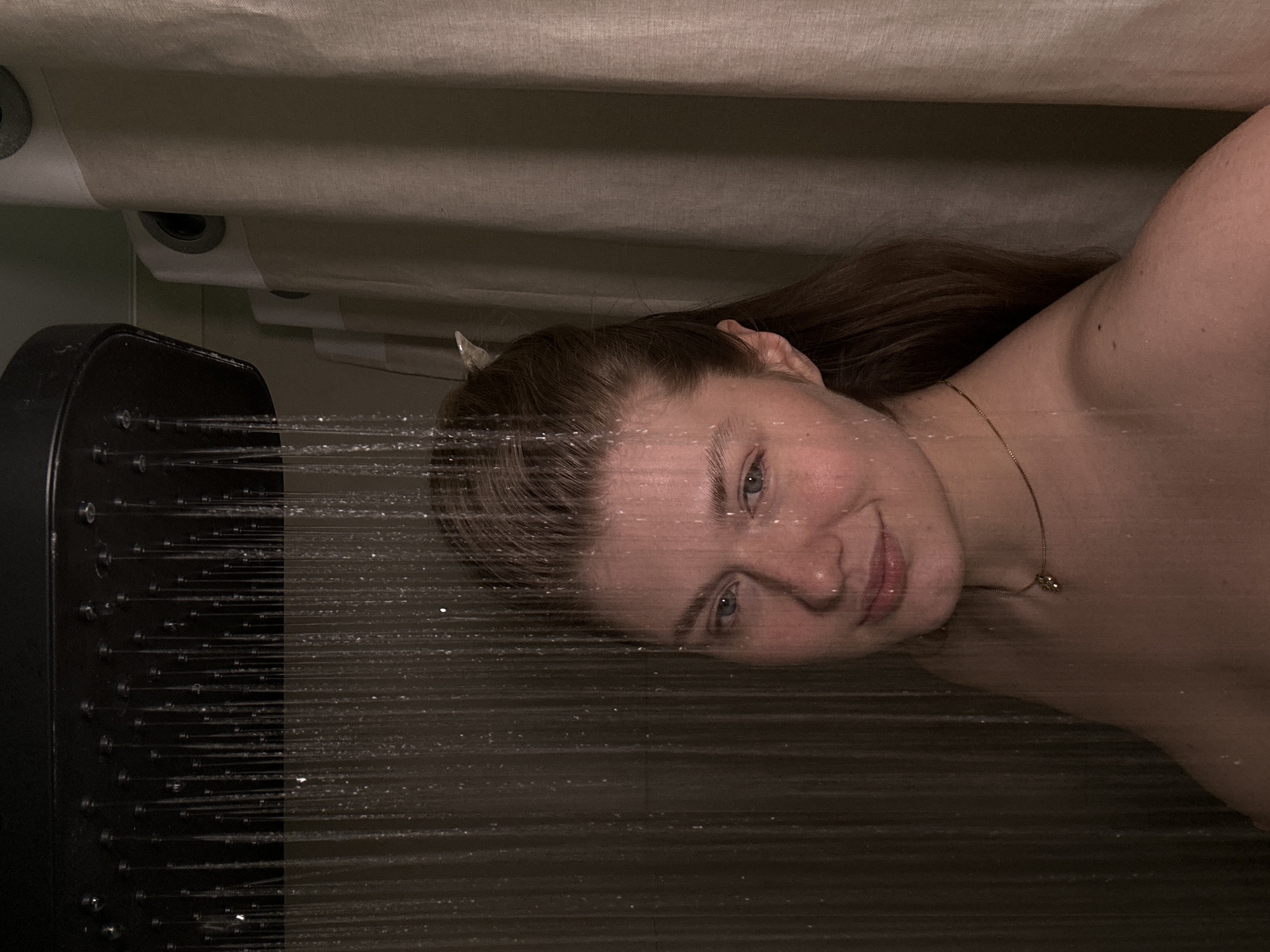A woman is taking a shower. The showerhead is a large, square, black fixture with multiple small holes, spraying water. The woman's face and upper body are visible through the water spray. She has fair skin, light eyes, and brown hair. A thin gold necklace is visible around her neck. A shower curtain is visible in the background. It is not possible to identify the specific location in Kazan, Russia from the image.