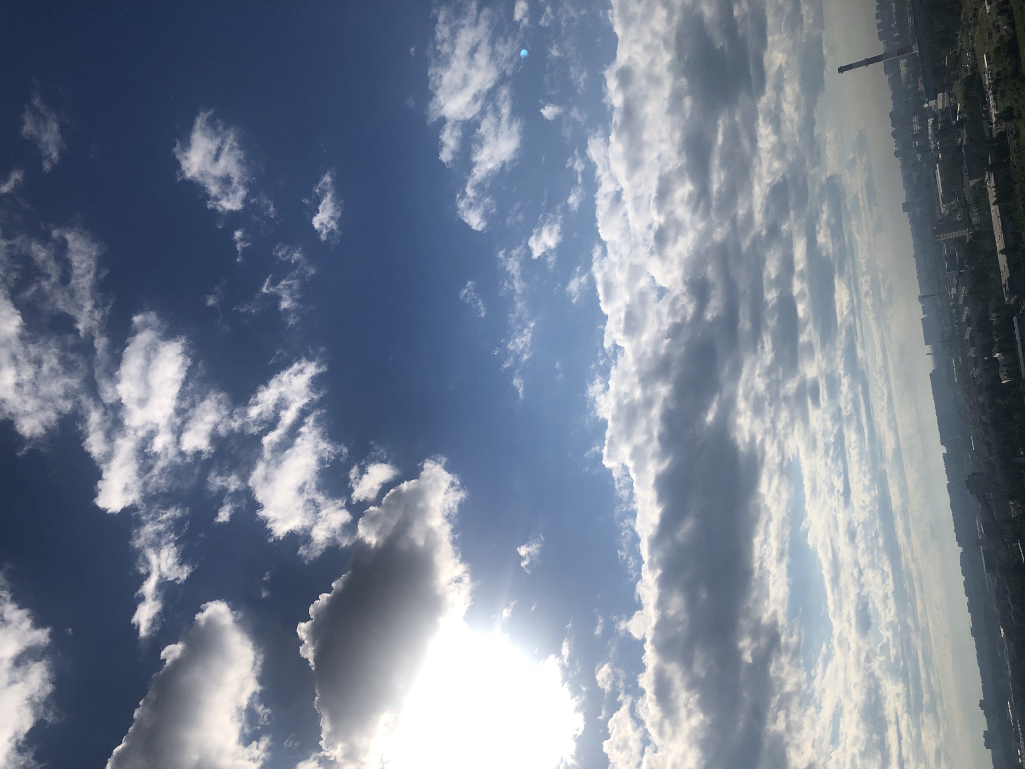 The image shows a wide shot of a cityscape under a bright, cloudy sky. The sun is partially obscured by a large, bright cloud formation in the upper left quadrant, casting rays of light. The sky is a vibrant blue with varying densities of white and gray clouds scattered throughout. The lower portion of the image reveals a dense urban landscape with numerous buildings, trees, and industrial structures. A prominent industrial smokestack is visible on the right side of the frame, with a hazy atmosphere present over the city. The perspective suggests the photo was taken from a high vantage point, possibly a tall building.