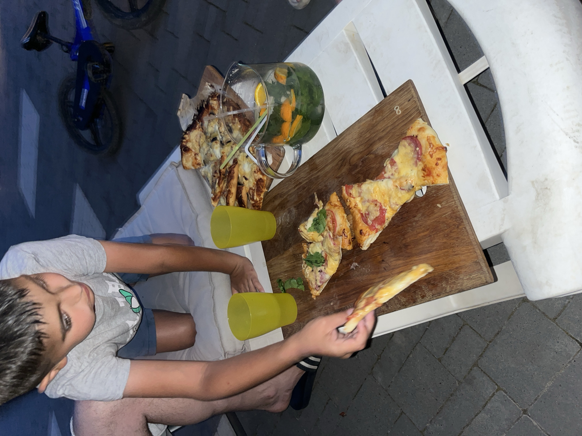 In Koroviia, Ukraine, a young boy in a grey t-shirt and blue shorts is on his knees on a white cushion, reaching for a slice of pizza from a wooden board. Next to the pizza are two yellow cups and a glass pitcher filled with a beverage containing citrus slices and mint. Behind him, a blue bicycle is partially visible. The scene appears to be outdoors, with a paved area in the background.