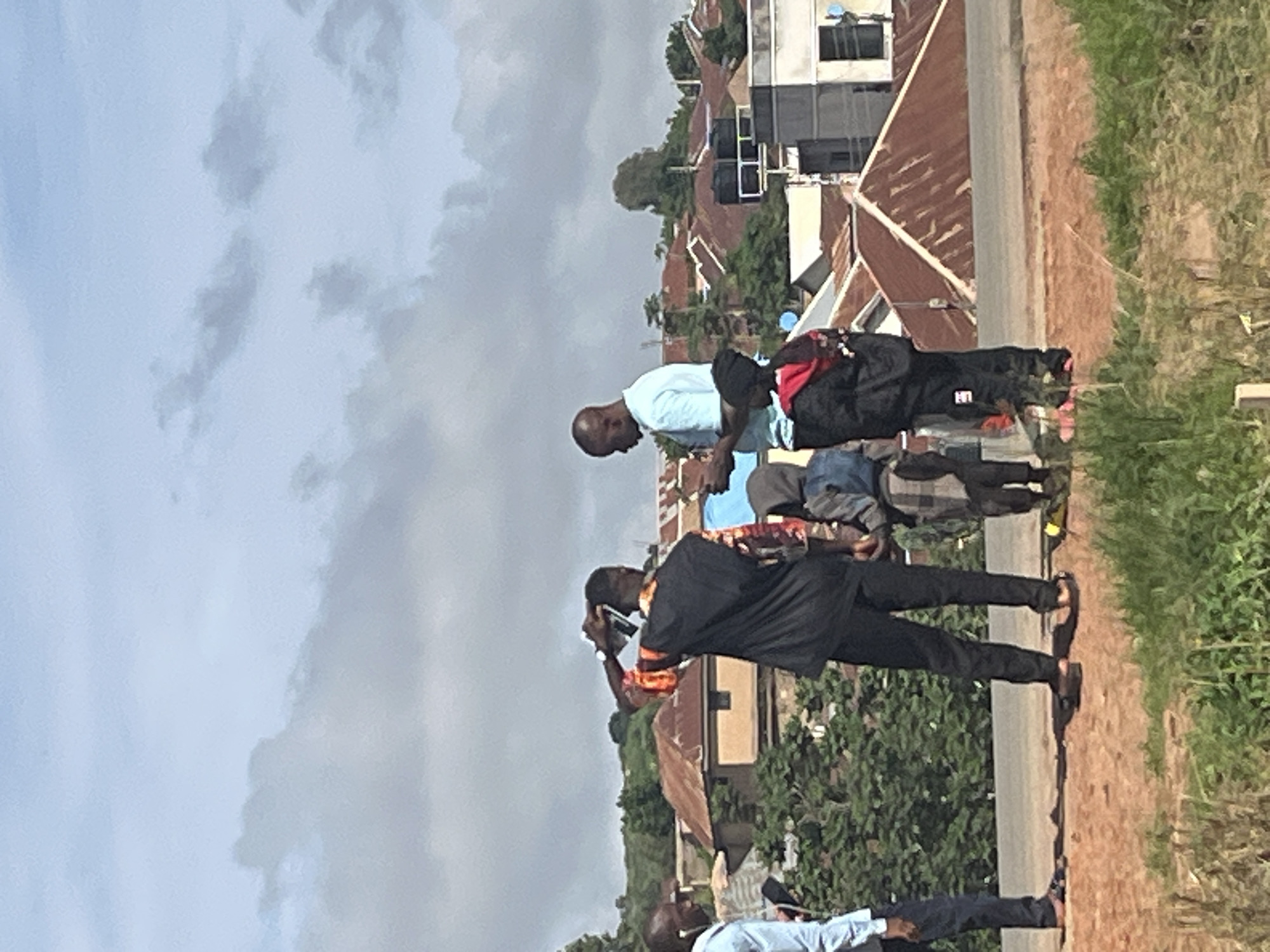 In Jos, Nigeria, on a sunny day with scattered clouds, a group of five individuals stands on an elevated dirt path, overlooking a densely populated area with numerous buildings featuring corrugated iron roofs. A man in a light blue t-shirt, holding a phone, is engaged in conversation with another man wearing a black garment over a patterned shirt. Behind them, three children, two boys and a girl, are also present. One boy wears a gray hoodie, while the other two, a boy and a girl, are dressed in a dark hooded jacket and a blue jacket, respectively. The environment appears to be a residential area on a hillside, with lush green trees and vegetation interspersed among the buildings.