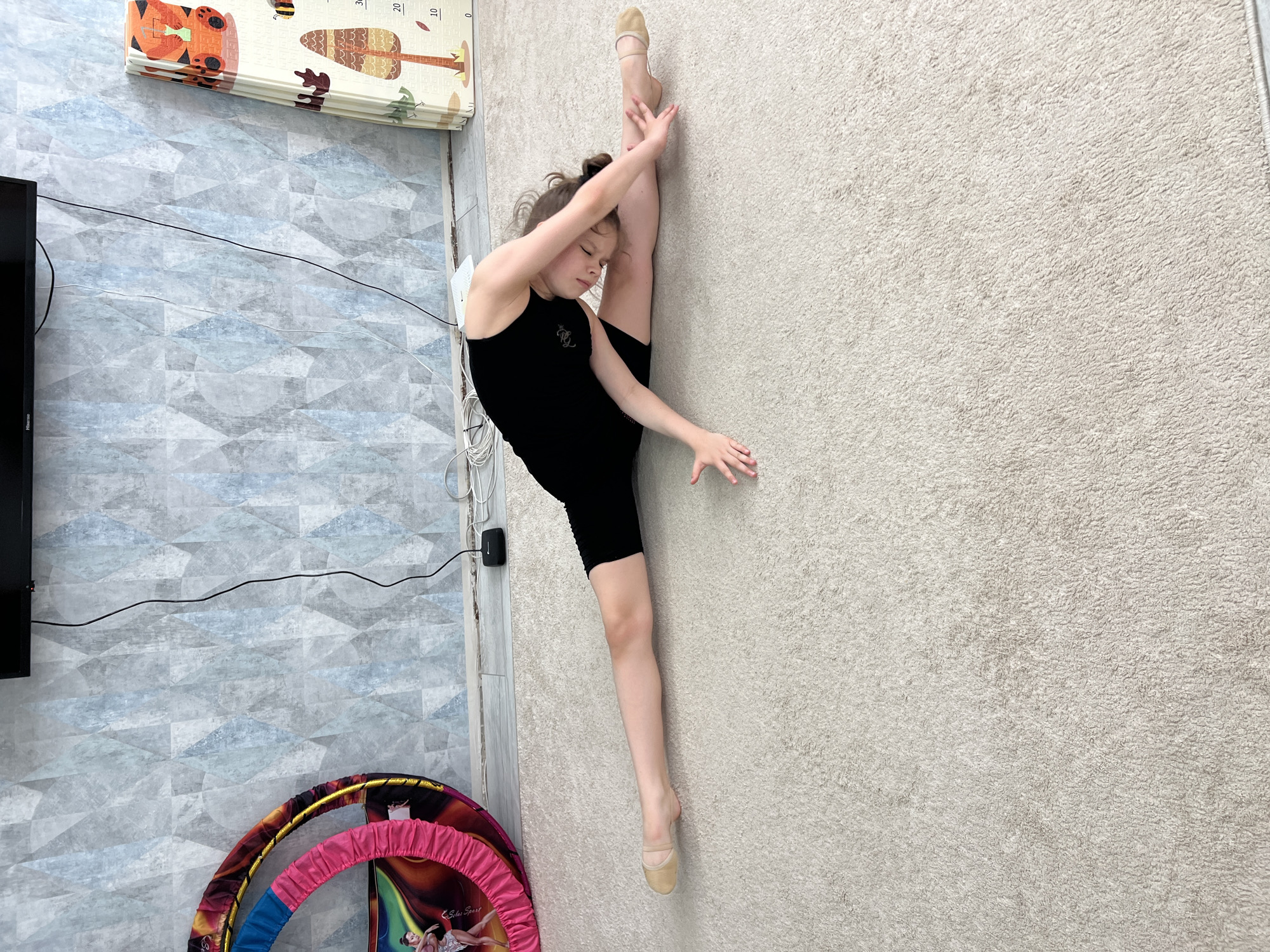 A young girl in a black leotard and tan ballet shoes is performing a vertical split against a light beige textured wall. She is holding her leg against the wall with her hand. Her other leg is extended down towards the floor. She has her hair pulled up in a bun. In the background, on a blue and grey patterned wall, there is a black television and a folded children's play mat with animal and tree designs. Below are several hula hoops. The scene appears to be indoors. The location is Chaikovsky, Russia.