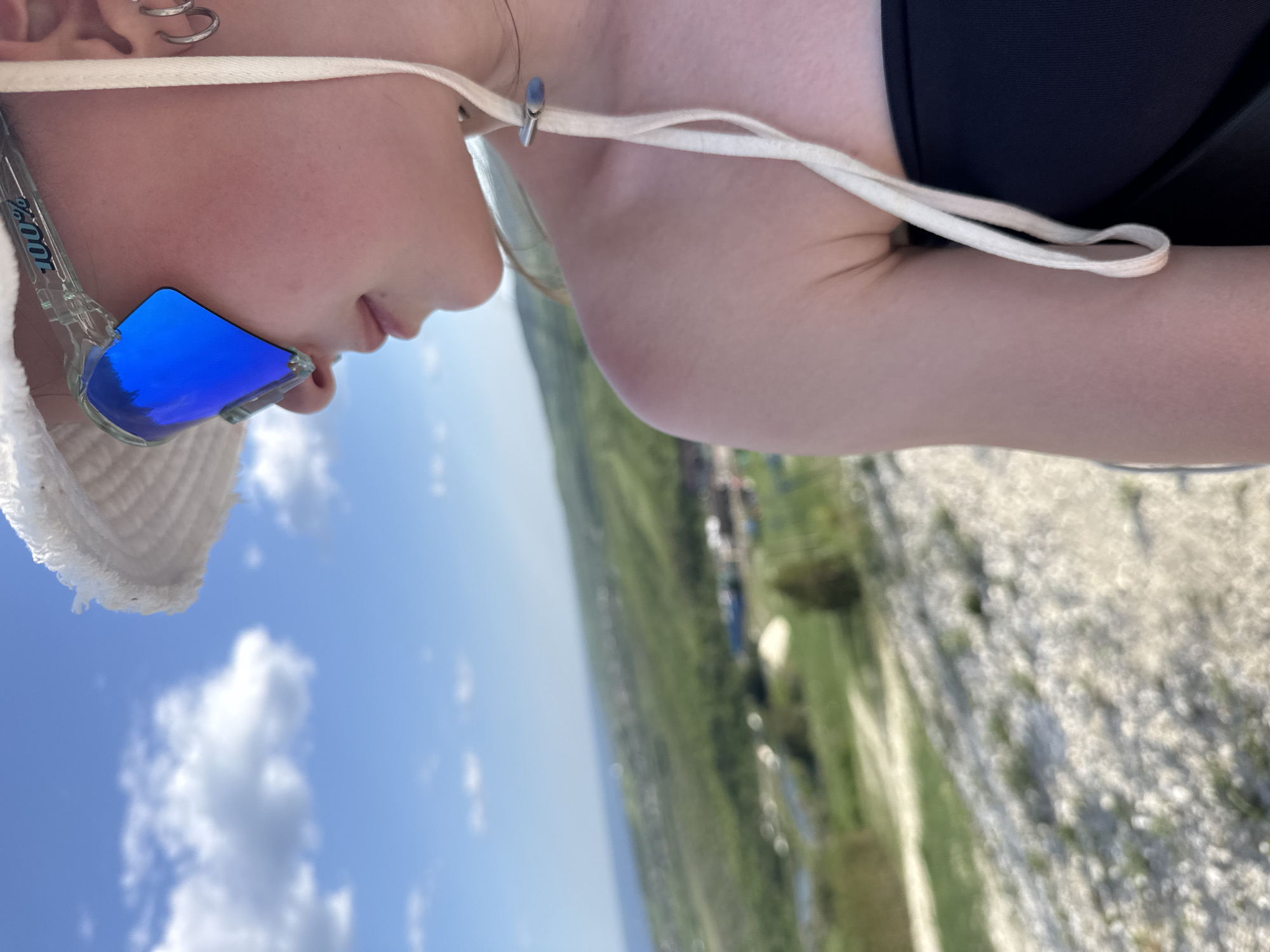 A close-up profile shot of a person with fair skin wearing sunglasses with reflective blue lenses. The person also wears a white hat with a fringed edge. The hat is secured with a white string. The background is an out-of-focus landscape of green fields and sky, likely taken in a rural or elevated location. A city is visible on the horizon, which could be Khvalynsk, Russia, given the request. The foreground contains a rocky, possibly limestone, surface. The photo appears to be taken outdoors during daylight hours.