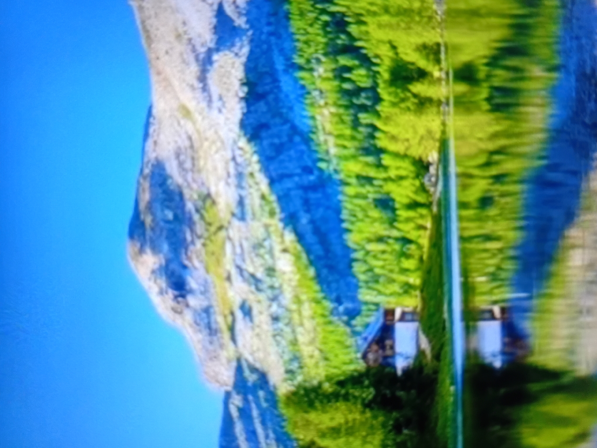 This media file depicts a serene mountainous landscape with a reflection in the foreground. The main subjects are a prominent rocky mountain on the left, covered in a mosaic of light and dark grey rock with patches of green vegetation, and a vibrant, lush green forest that cascades down the mountain's slopes. In the distance, nestled within the forest, are a couple of small, rustic-looking houses with dark roofs. The sky is a clear, bright blue, suggesting a sunny day.
The scene is a mirror image, with the mountain, forest, and houses reflected in a body of water that dominates the lower half of the frame. The reflection is slightly distorted, creating a wavy effect, but clearly shows the contours of the mountain and the dense greenery of the forest. The water itself appears calm, contributing to the peaceful atmosphere.
There are no people visible in the media file. The overall impression is one of natural beauty and tranquility, possibly during daylight hours given the brightness of the sky and the clear visibility of the landscape. The presence of the houses hints at a remote, possibly rural or alpine settlement. The image lacks any visible text. The scene evokes a sense of calm and immersion in nature.